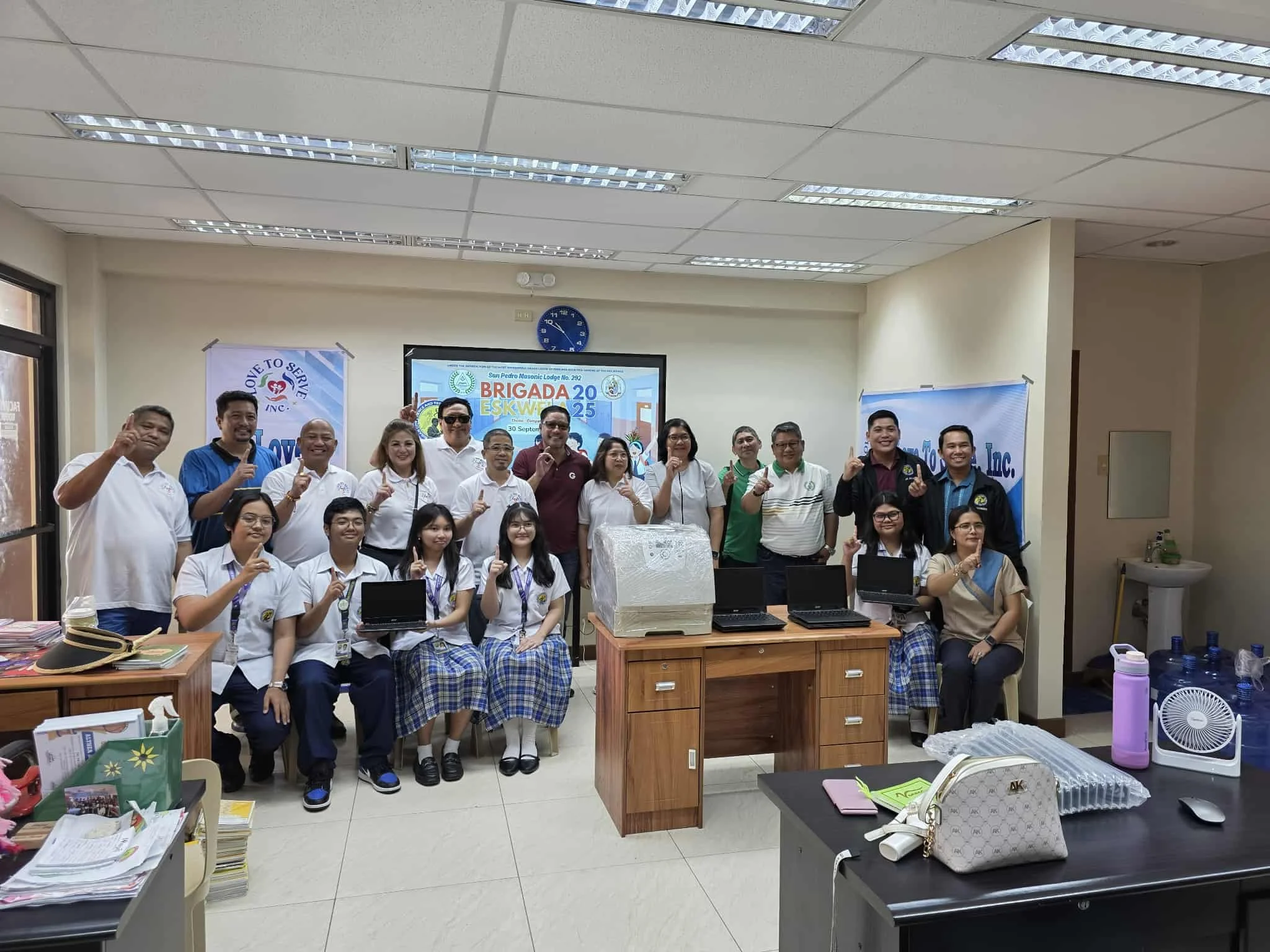 A group of students and adults posing for a photo in a classroom or conference room. There are laptops and various items on desks in the foreground. The people are making a gesture with their hands, and a digital screen behind them displays informati