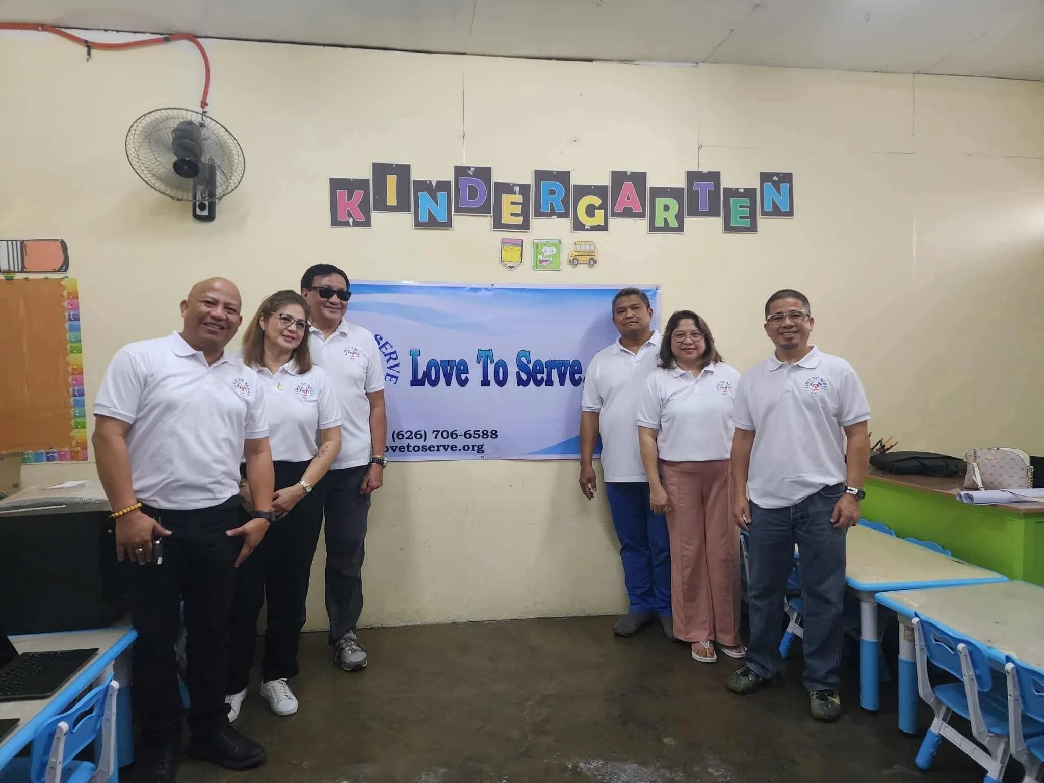 Group of six adults standing in a kindergarten classroom in front of a banner that reads 'Love To Serve' with additional text and a website link. They are wearing white polo shirts with a logo, smiling at the camera.