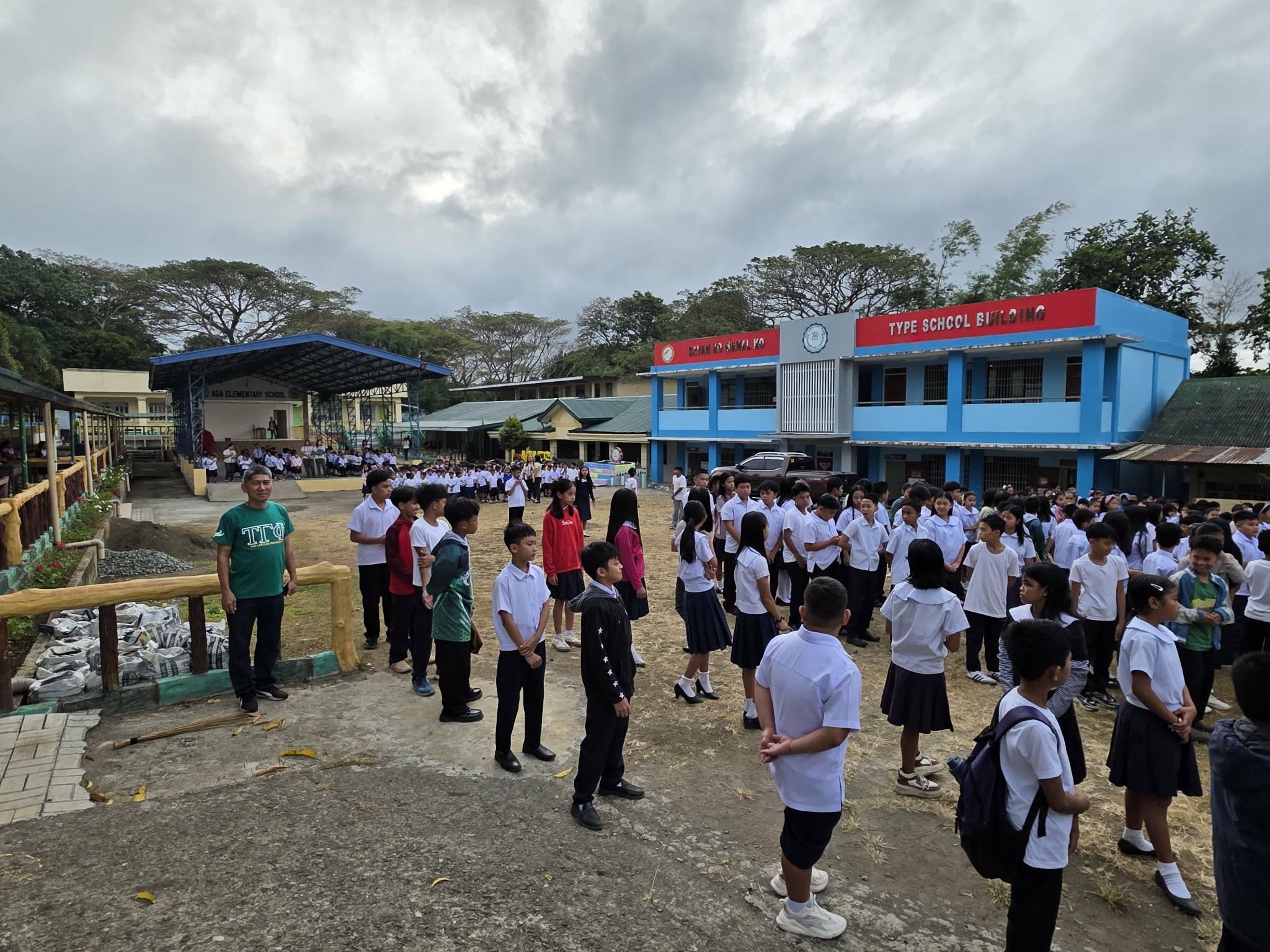 Students gathered outside a school building during a morning assembly, with many students in uniform standing in formation and a stage on the left side where some students are performing or speaking.