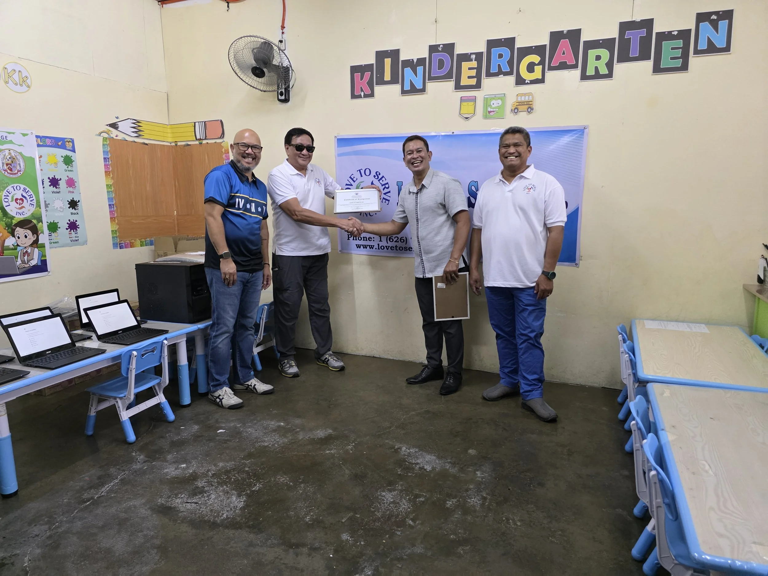 Four men standing inside a kindergarten classroom, with one man handing a document or check to another. The classroom has blue tables and chairs, computers, and colorful decorations, including a sign that says Kindergarten.