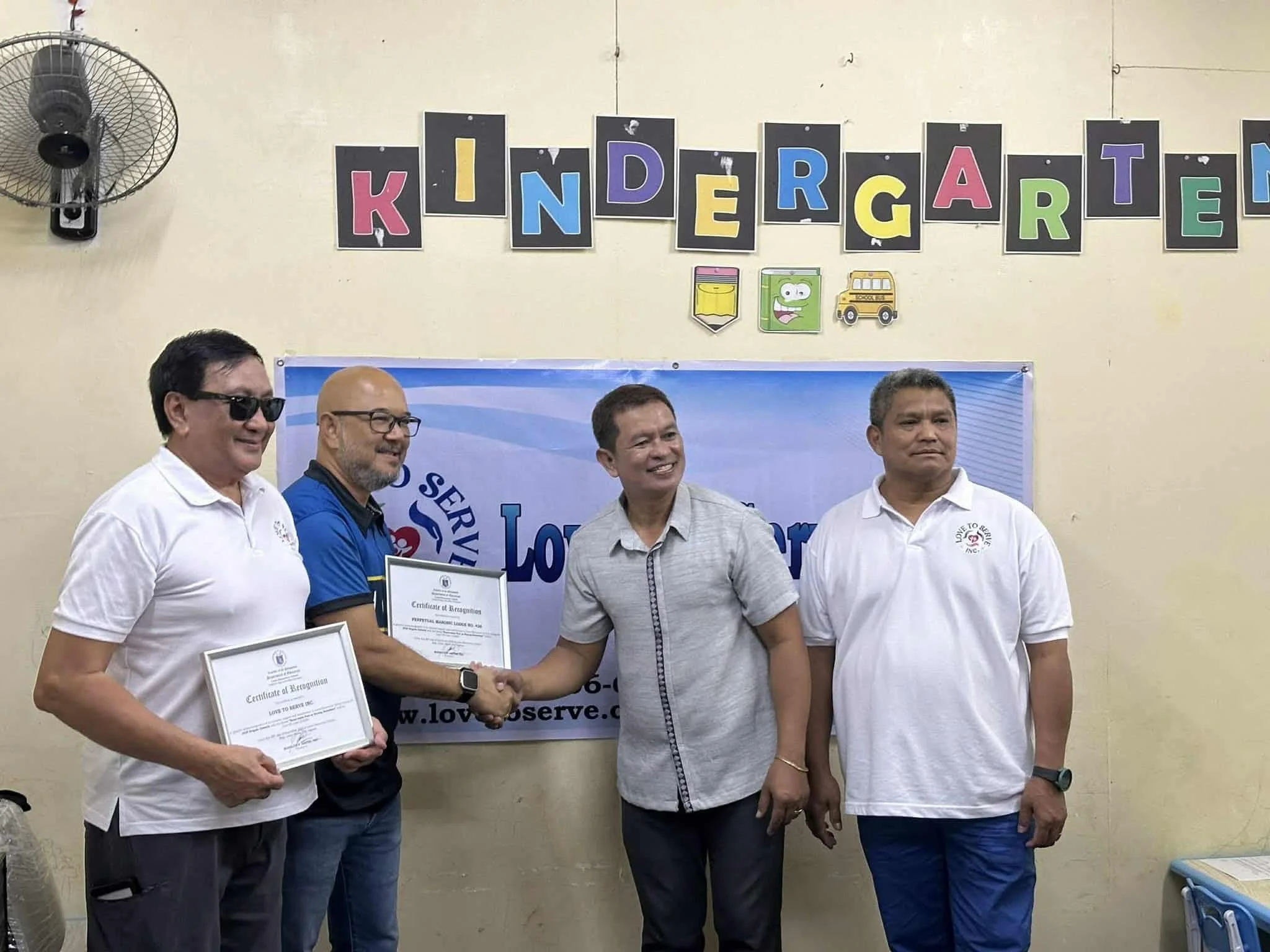 Four men standing in front of a wall with a 'Kindergarten' banner, two holding certificates, one shaking hands, all smiling