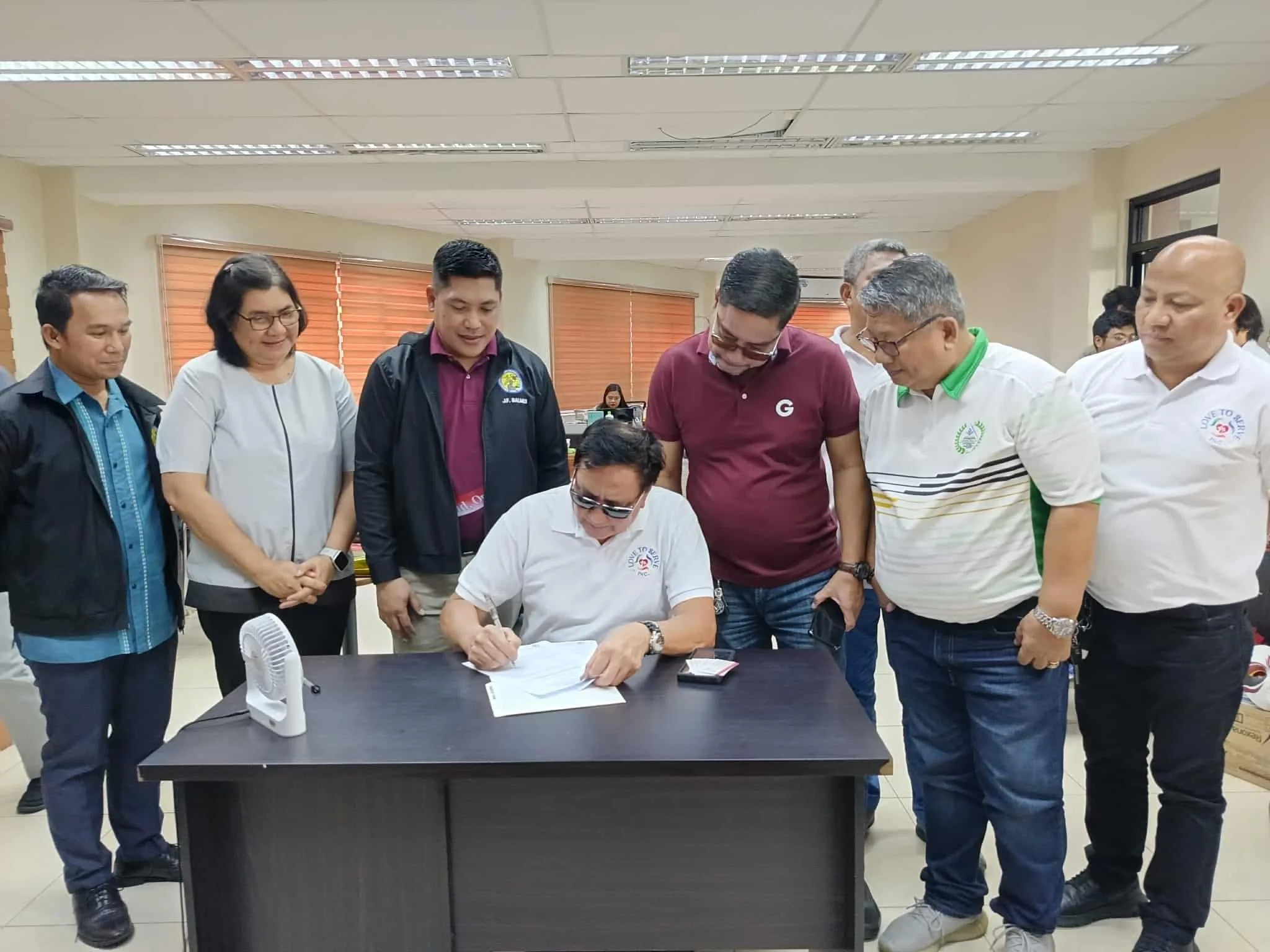 A group of people gathered around a man sitting at a desk, signing papers. The man appears to be in an official setting, with some people watching and others observing the signing.