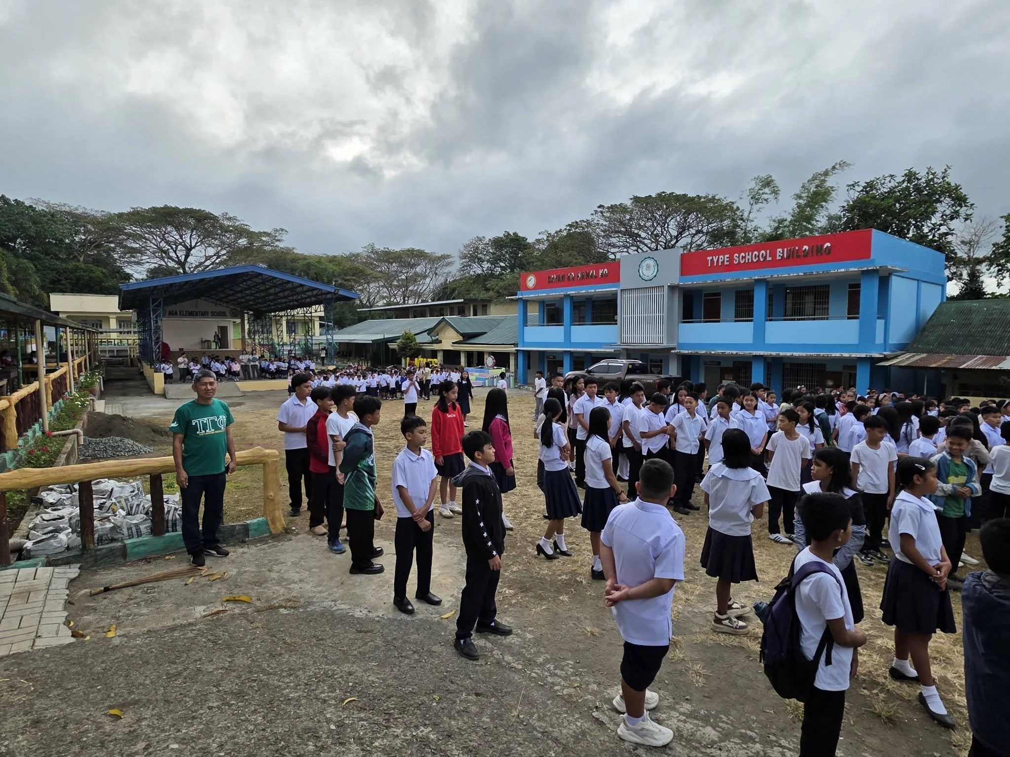 Students gathered in the school yard for an event at Aga Elementary School, with a stage and a blue school building in the background, surrounded by trees.