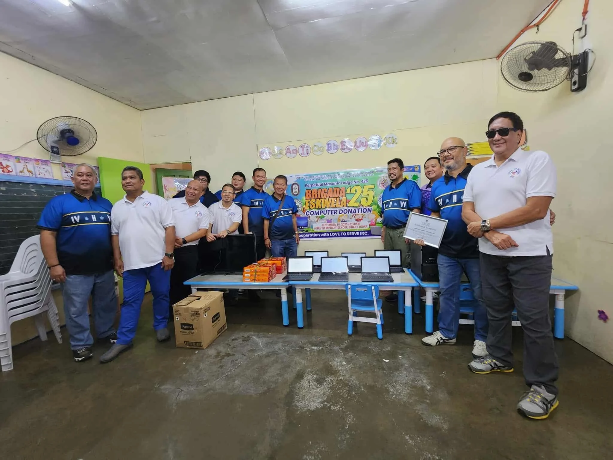 Group of men standing in a classroom during a computer donation event, with laptops on tables and a banner in the background.