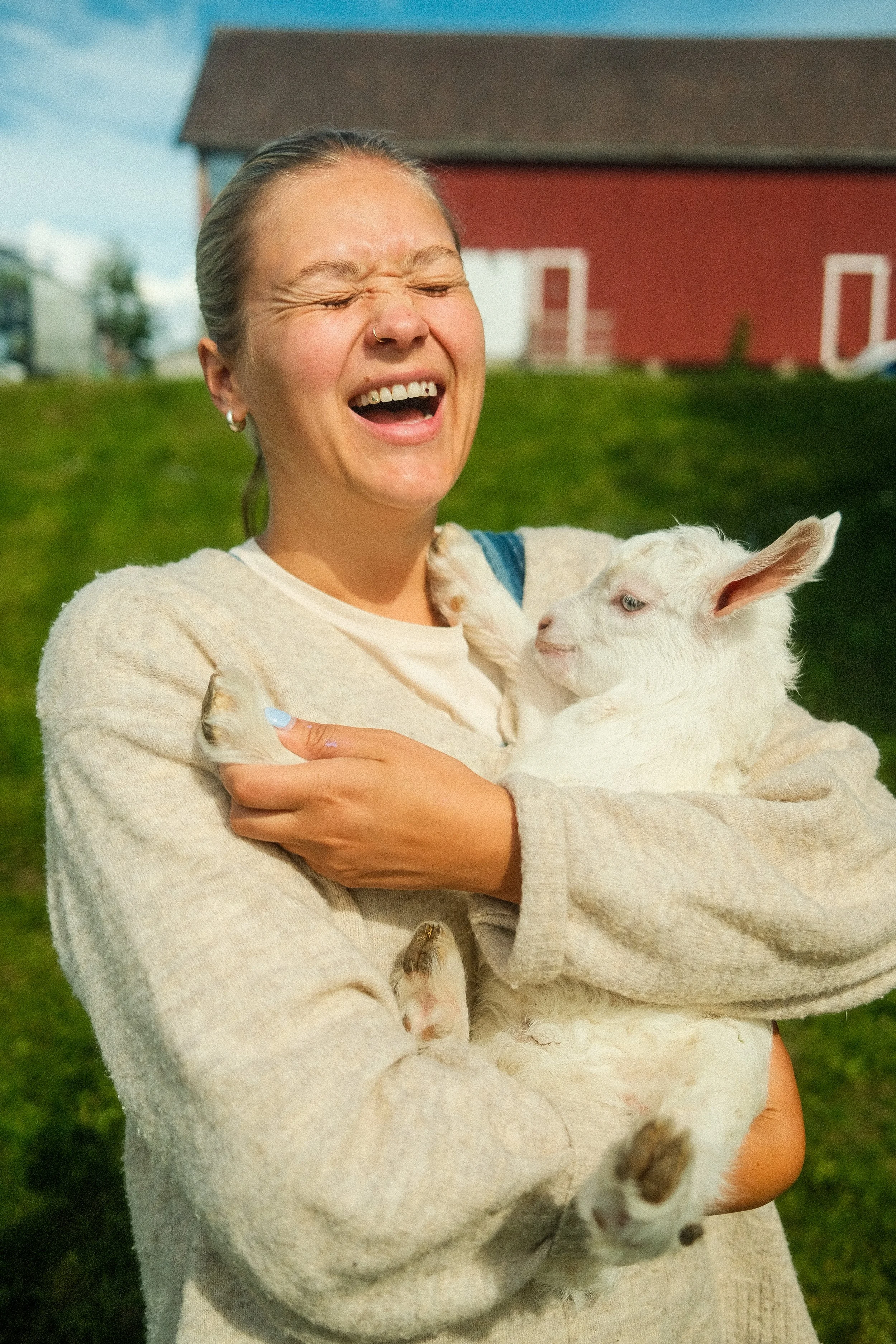 A woman smiling and laughing while holding a white baby goat outdoors on a farm with a red barn in the background.