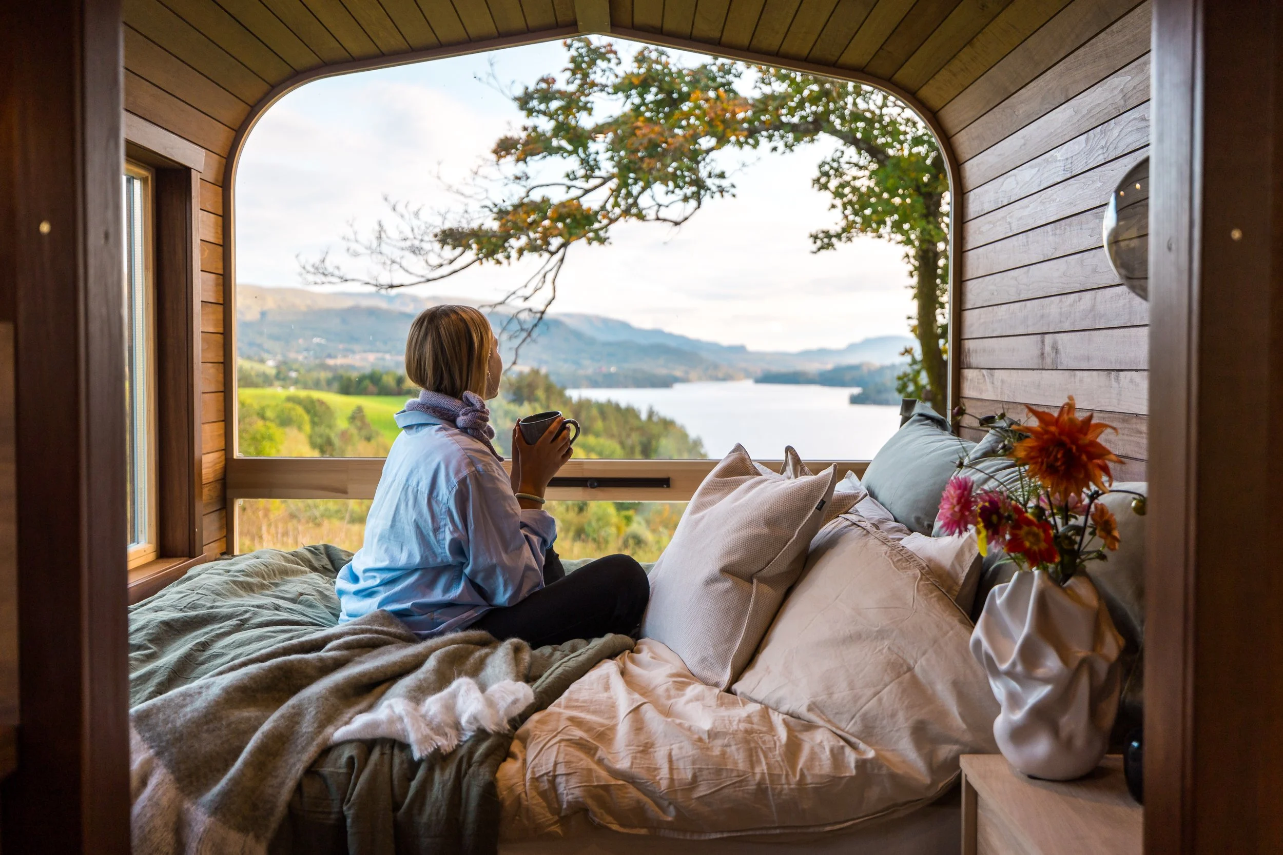 Woman sitting on bed in a cozy wooden cabin, holding a mug, looking out at a scenic landscape with a river, trees, and mountains.