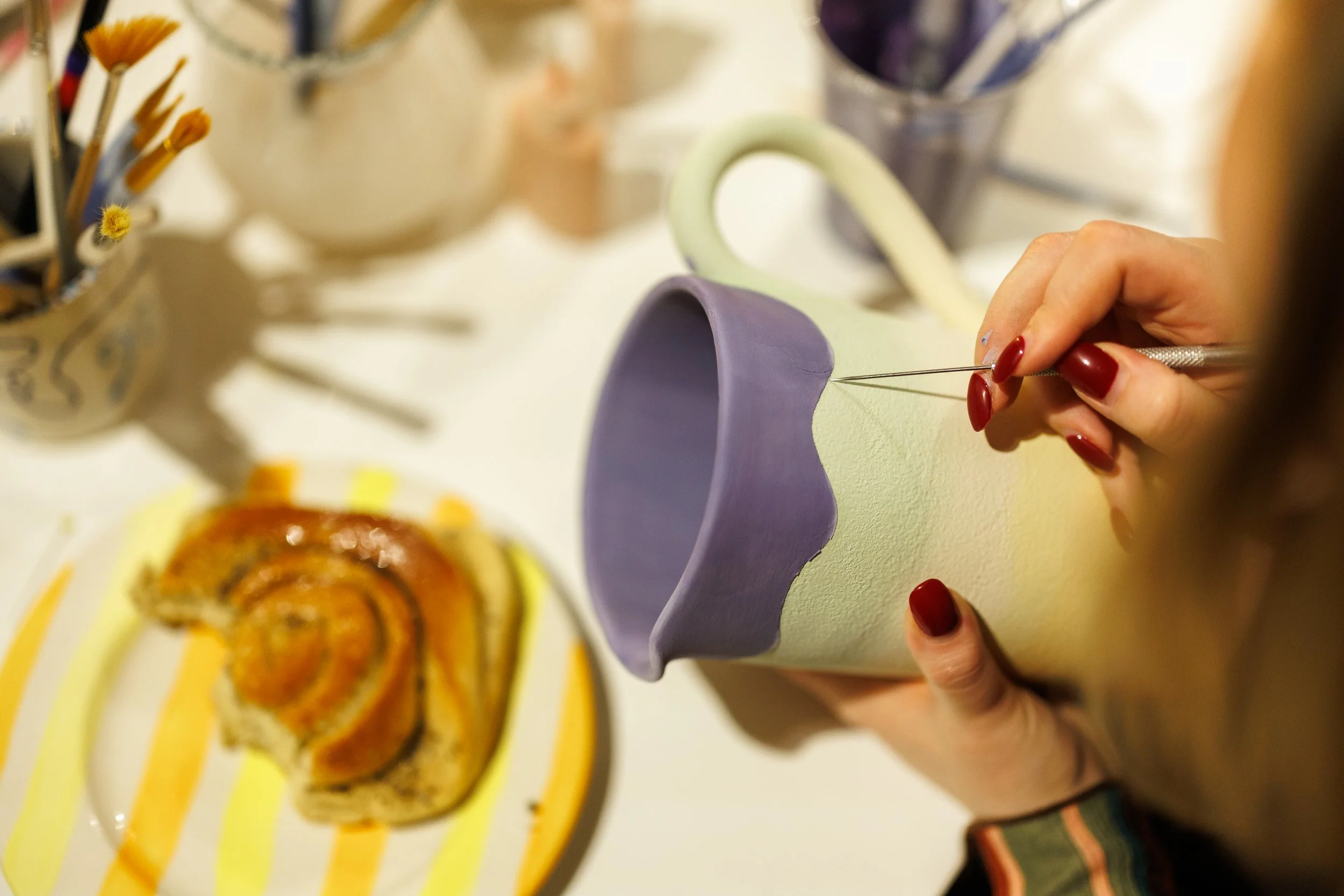 A person using a fine tool to decorate a pastel-colored ceramic mug, with a plate of pastry and a breakfast-themed table setting visible in the background.