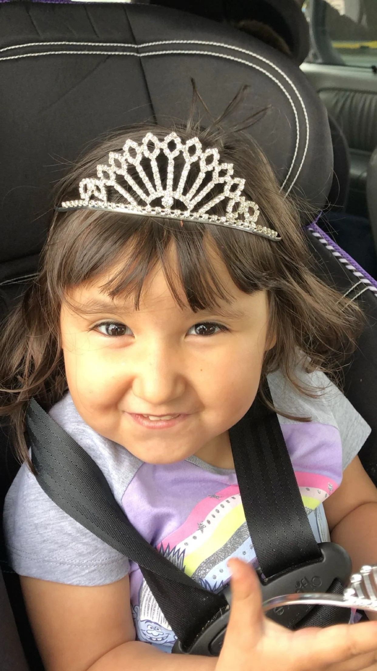 A young girl sitting in a car seat, wearing a small tiara and smiling.