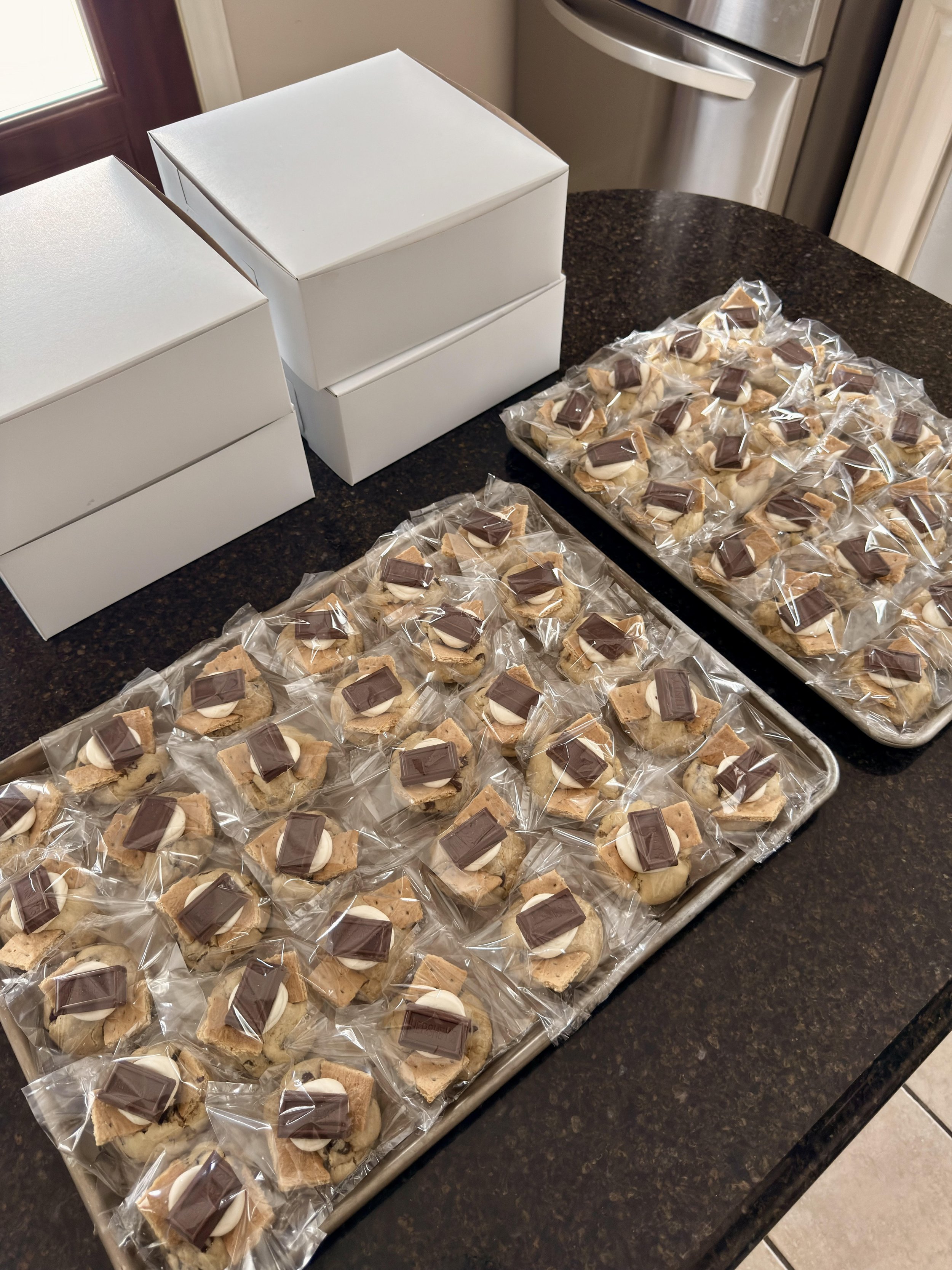 Tray of small desserts topped with chocolate squares, individually wrapped in clear plastic on a kitchen countertop.