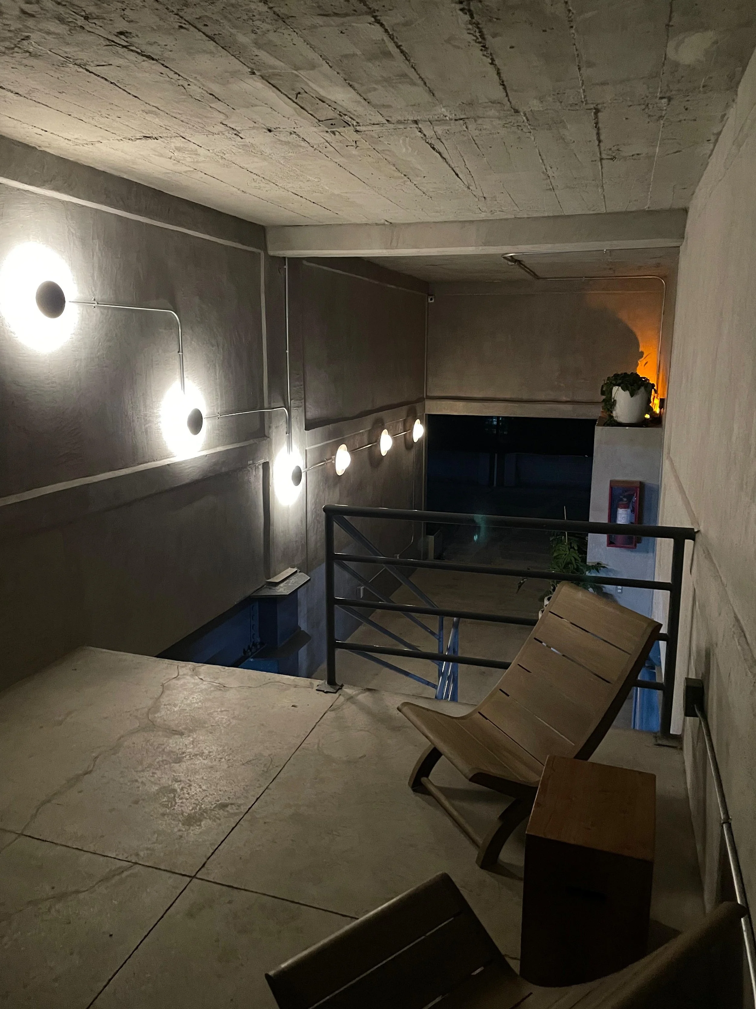 An indoor space with a concrete ceiling and floor, wall-mounted lights, wooden chairs, a railing, potted plants, and a fire extinguisher.