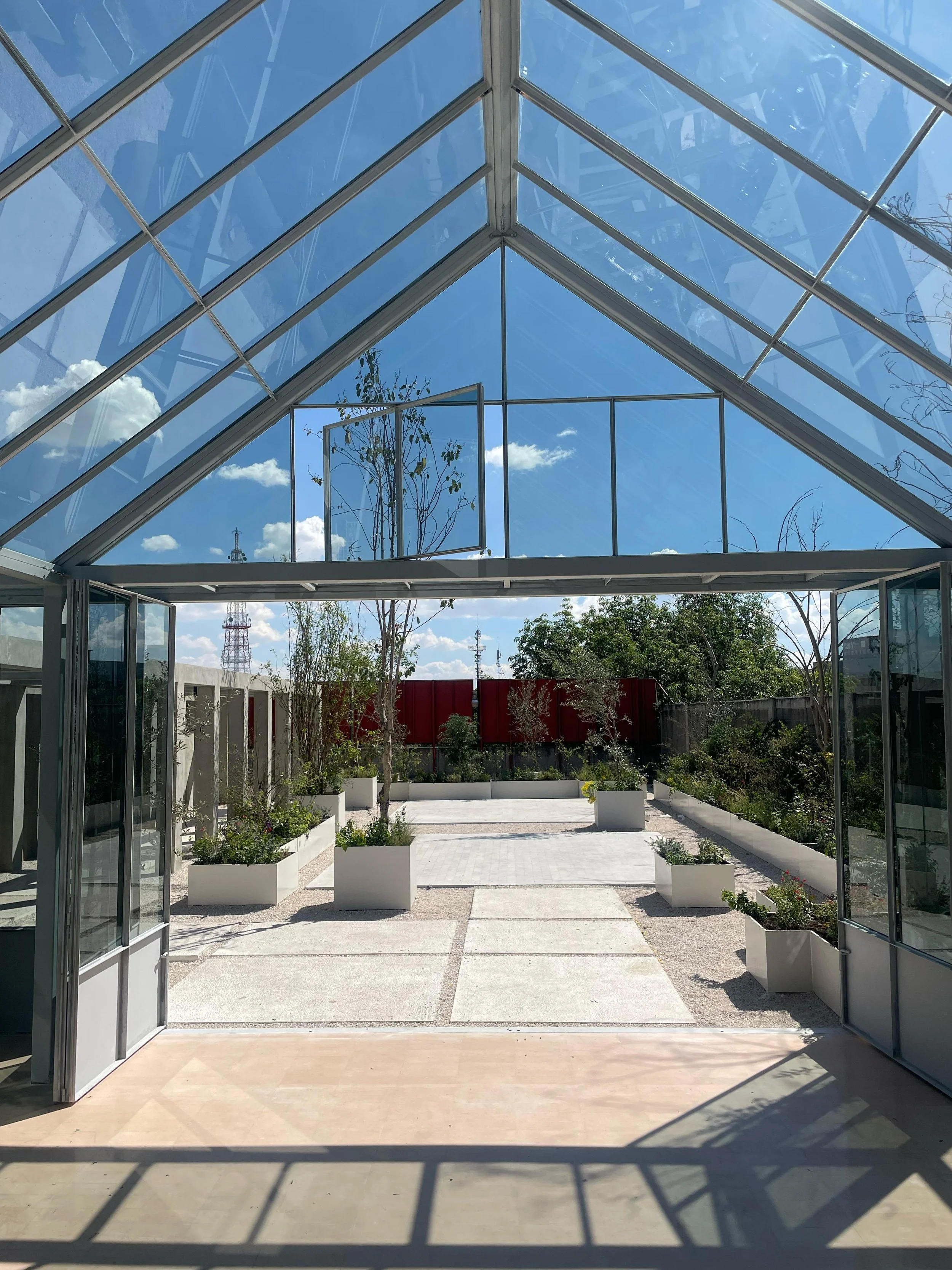 View from inside a glass greenhouse looking out onto a garden with potted plants, trees, and a blue sky with clouds.