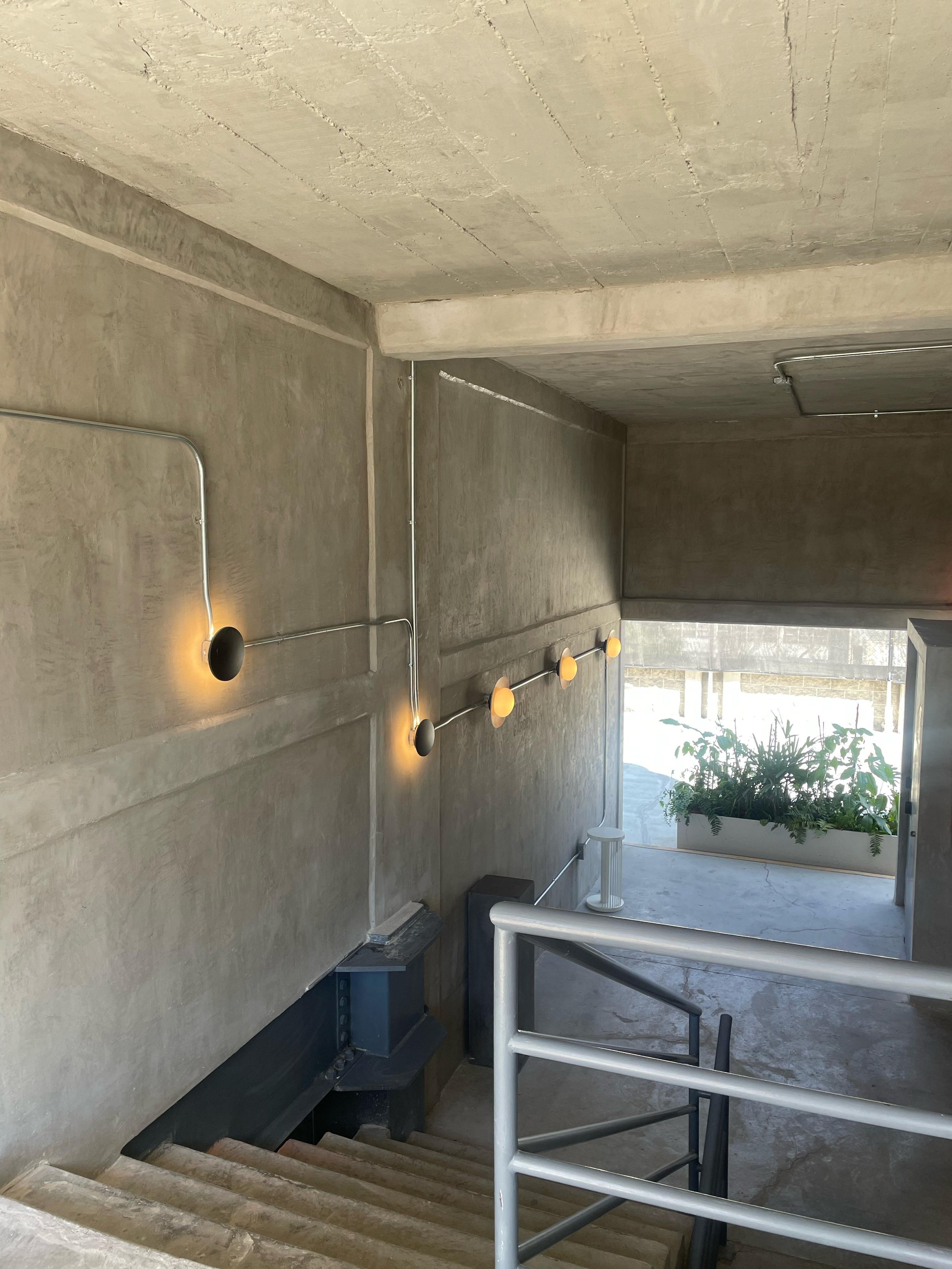 Concrete staircase leading down with a metal railing, gray concrete walls and ceiling, wall-mounted lights, and a window with a planter box of greenery outside.