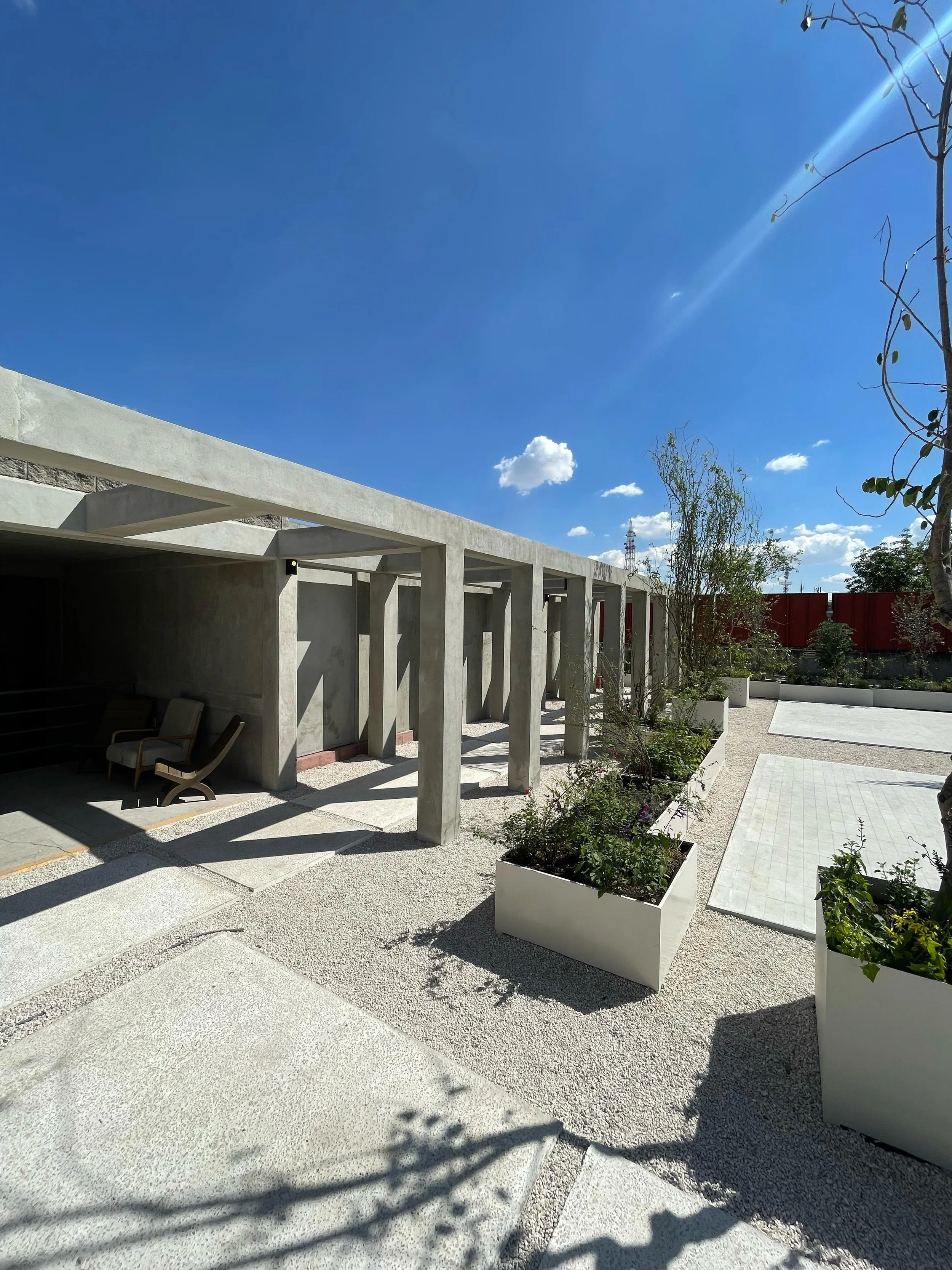 Modern outdoor patio area with concrete structures, potted plants, and seating, under a blue sky with a few clouds.