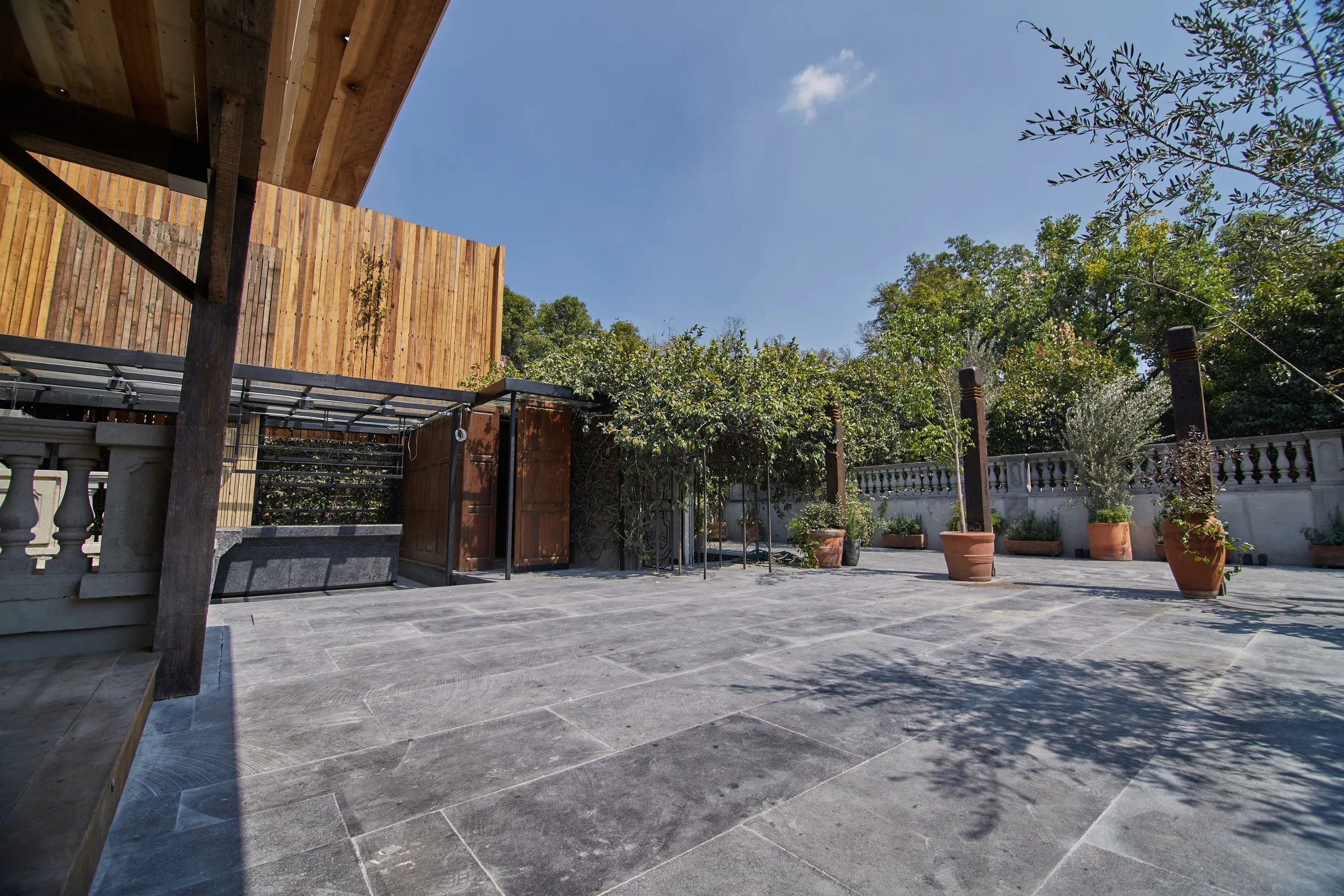 Outdoor patio with gray stone tiles, potted plants, trees, and a white stone wall with balustrade.