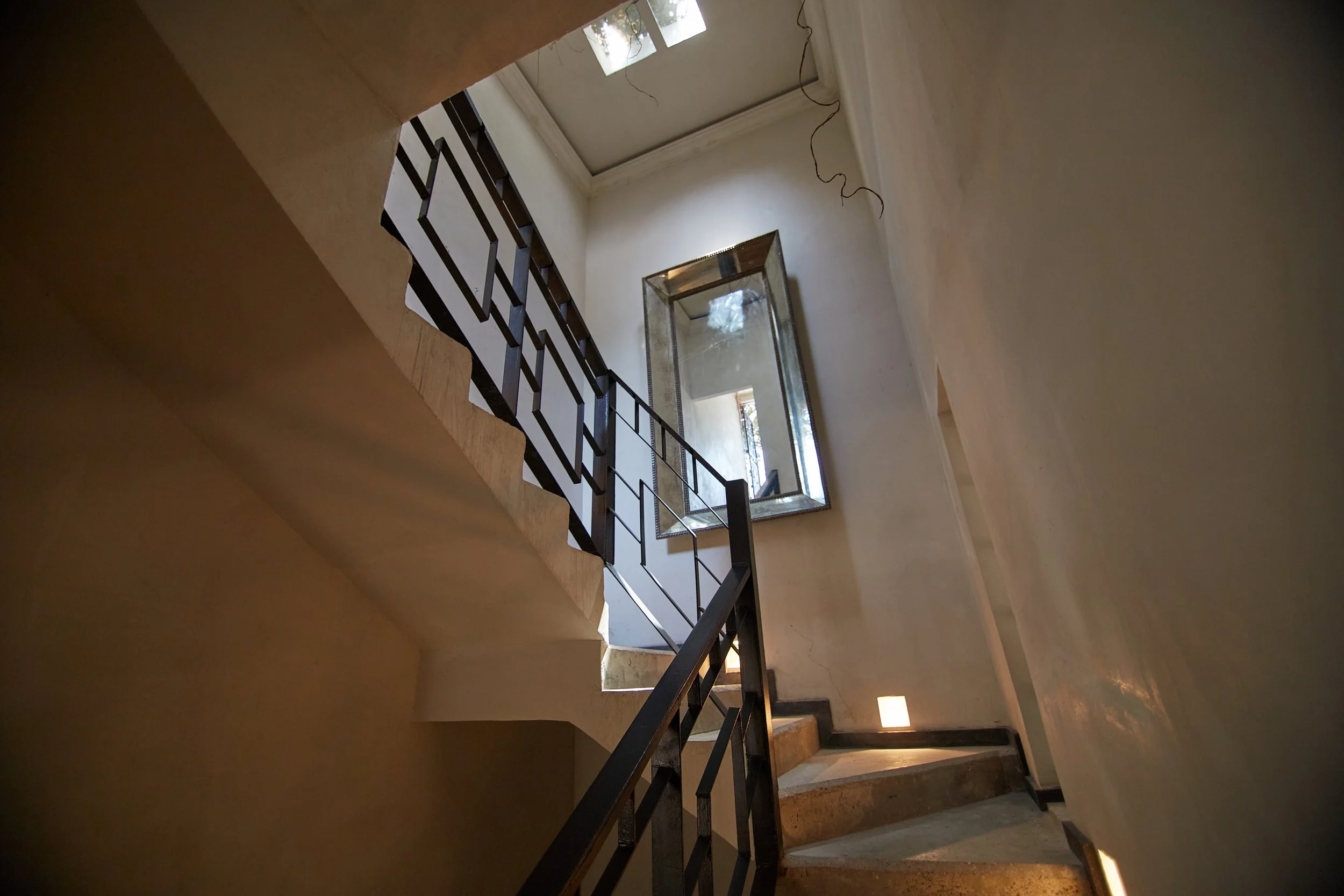 Indoor staircase with black metal railing, beige steps, a large rectangular mirror on the wall, and natural light coming from a window at the top.