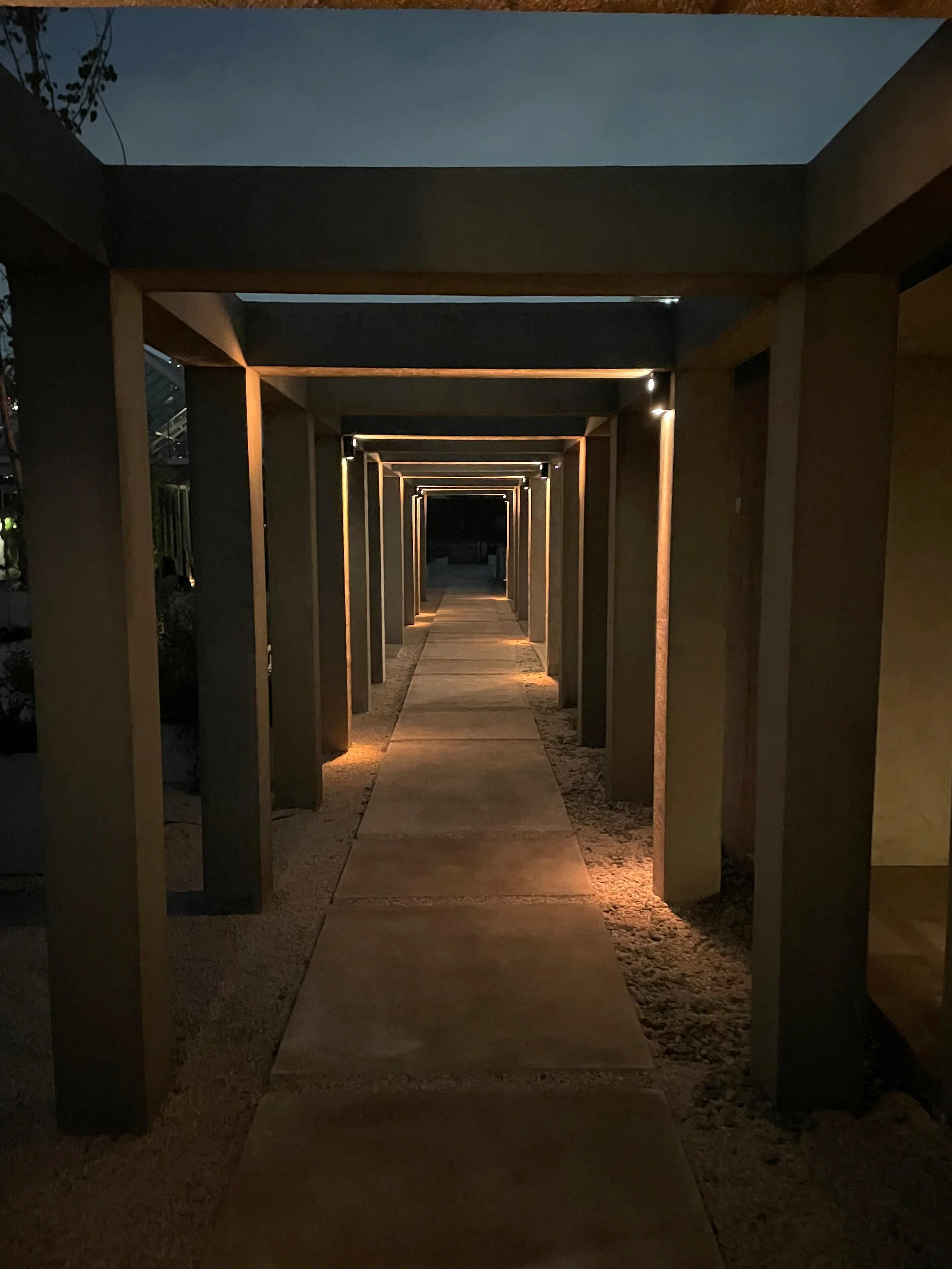 A covered outdoor walkway at night with warm lighting along the edges of the columns, creating a repetitive pattern of rectangular frames.