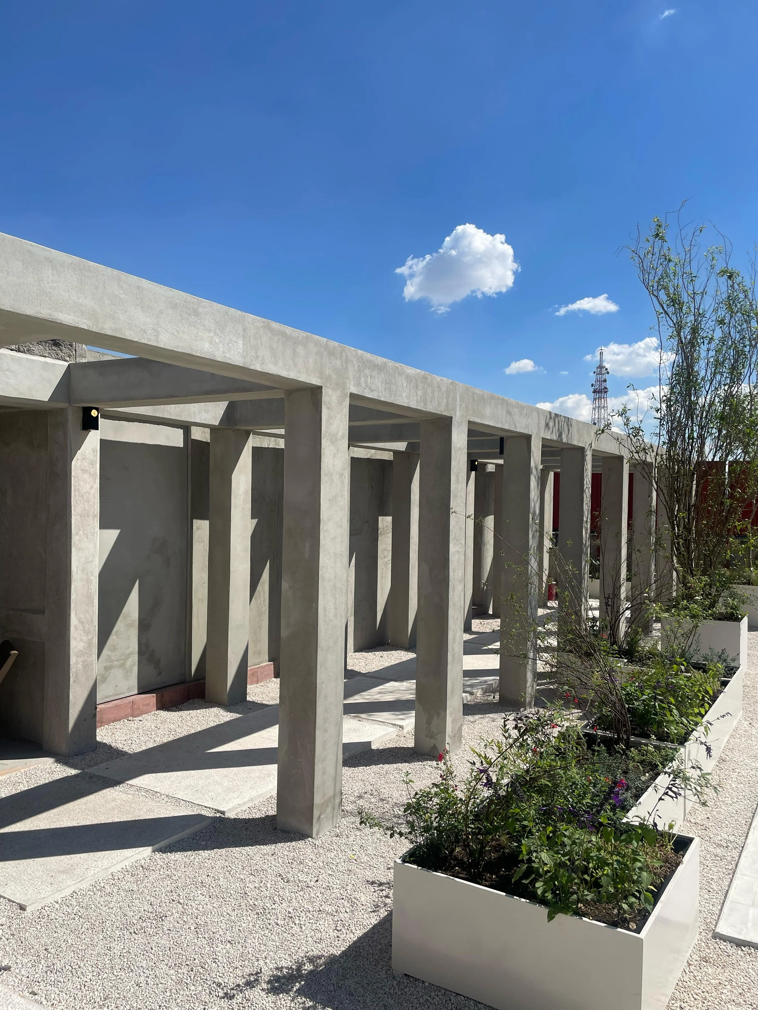 Modern building exterior with concrete pillars and planters with plants, under a blue sky with white clouds.