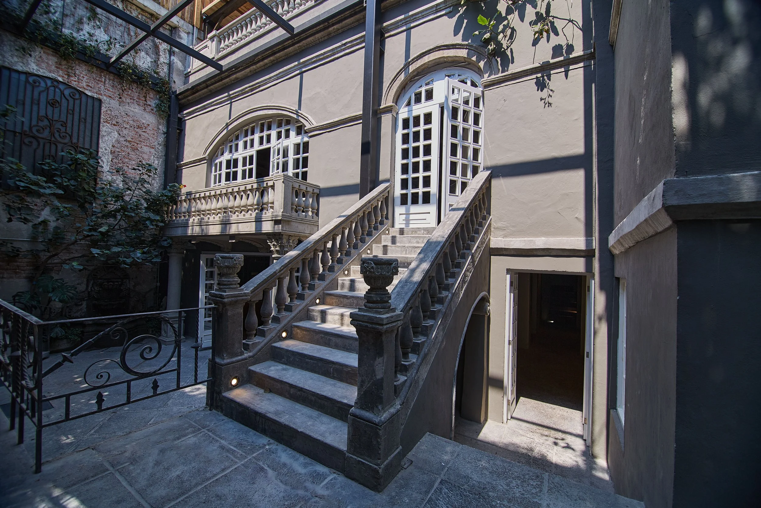 Stone staircase leading to a white door with an arched window and decorative railing, adjacent to a building exterior with a balcony and plants