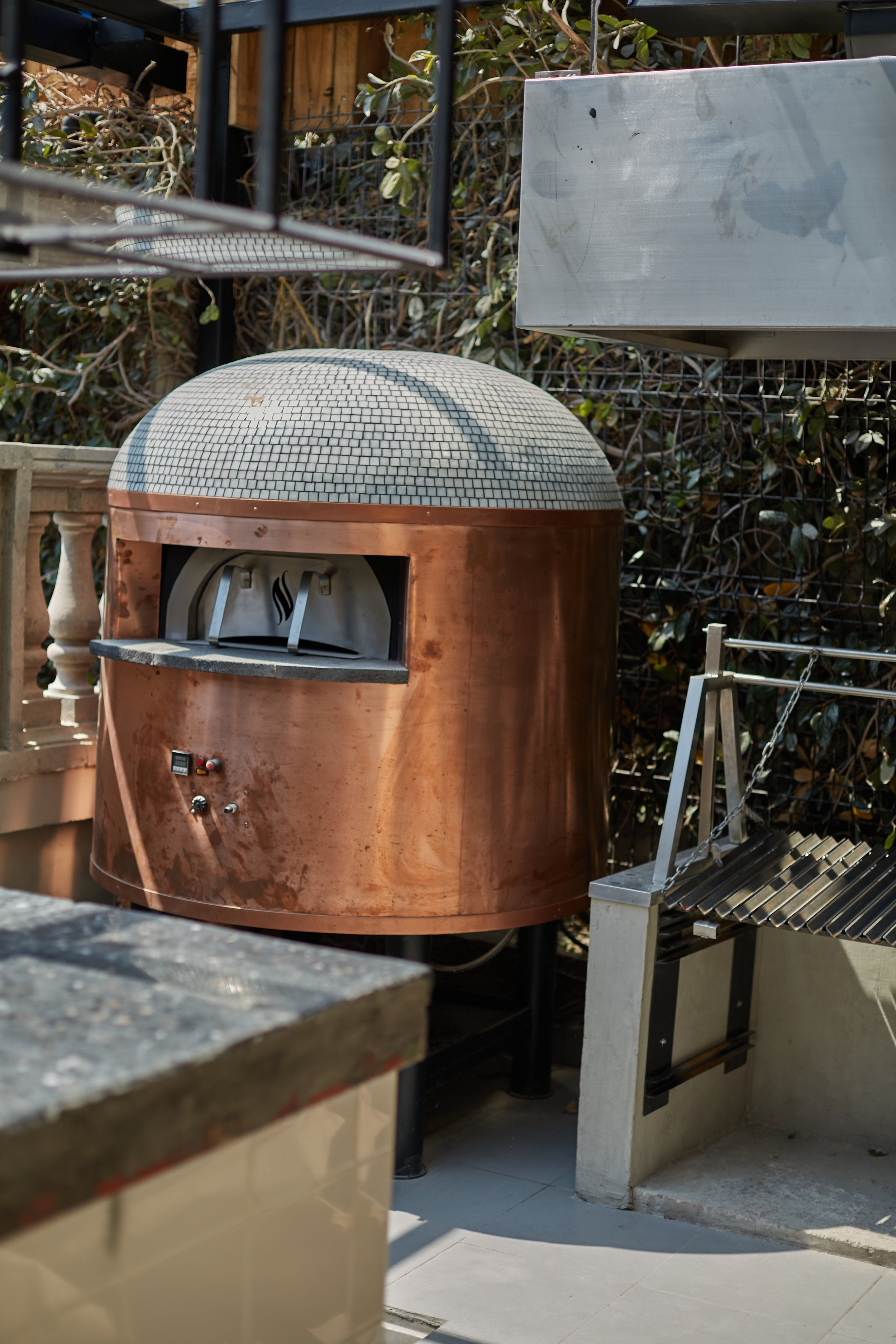 A copper-colored outdoor pizza oven with a mosaic dome, situated on a patio area with various metal and stone furnishings.