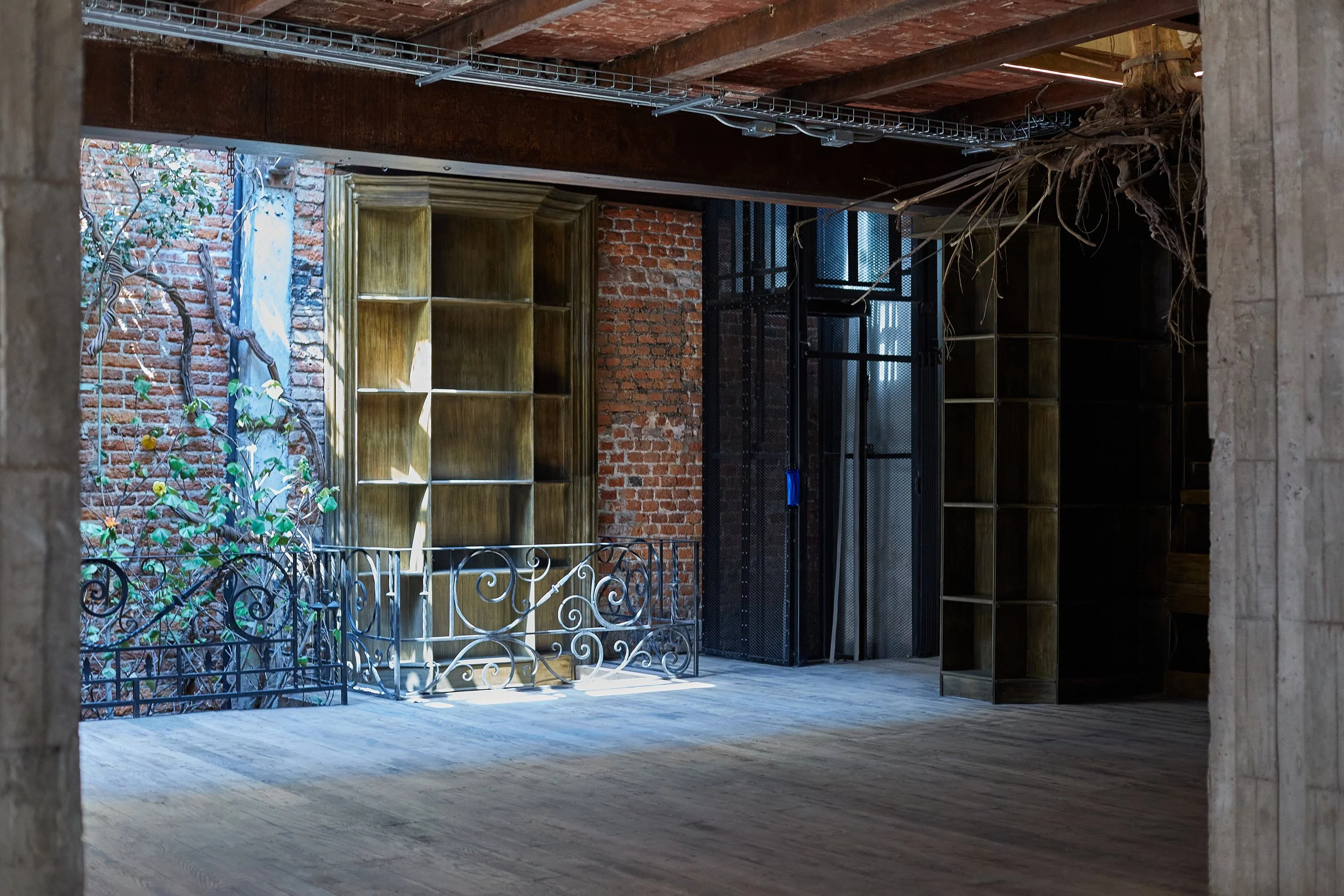 Interior of a building with exposed brick walls, wooden ceiling beams, empty wooden bookshelves, metal railing, and a metal elevator door.
