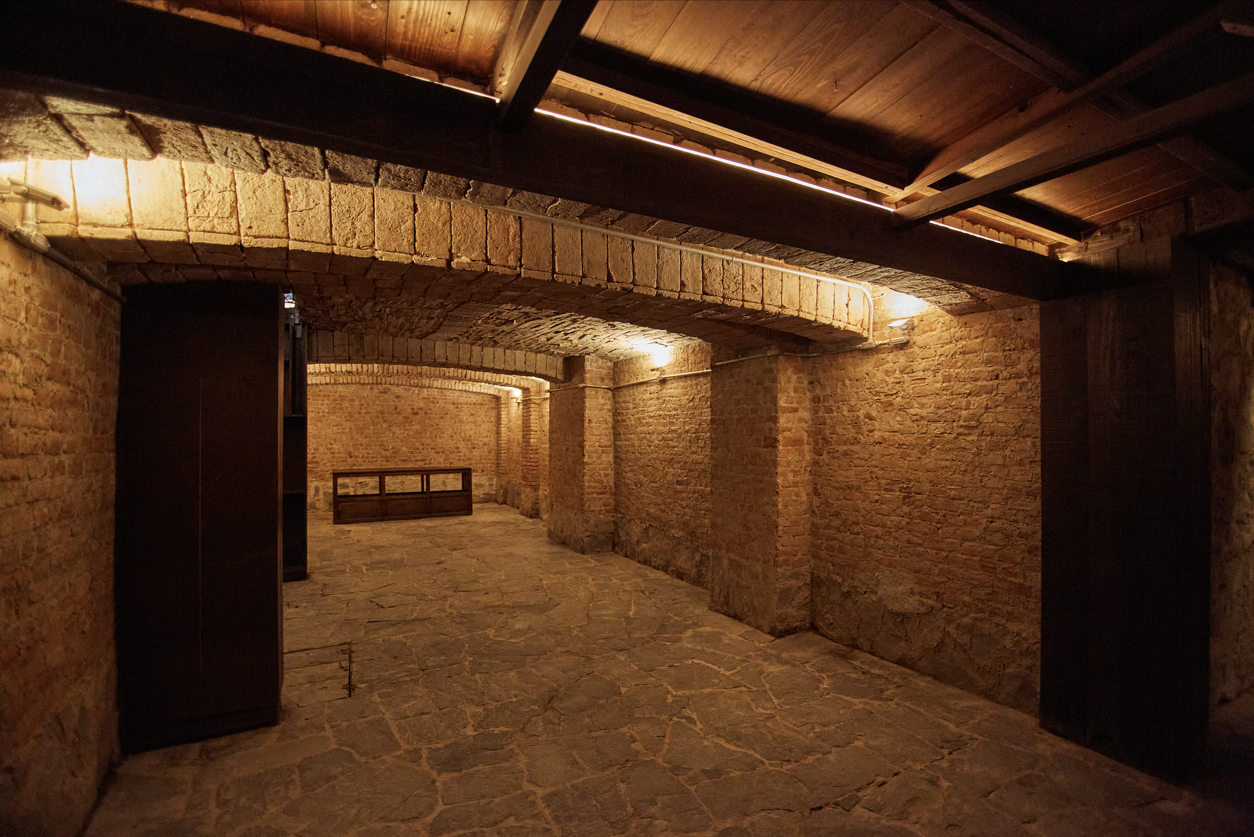 Underground brick brick-walled cellar with stone floor and wooden ceiling, illuminated by warm lights.