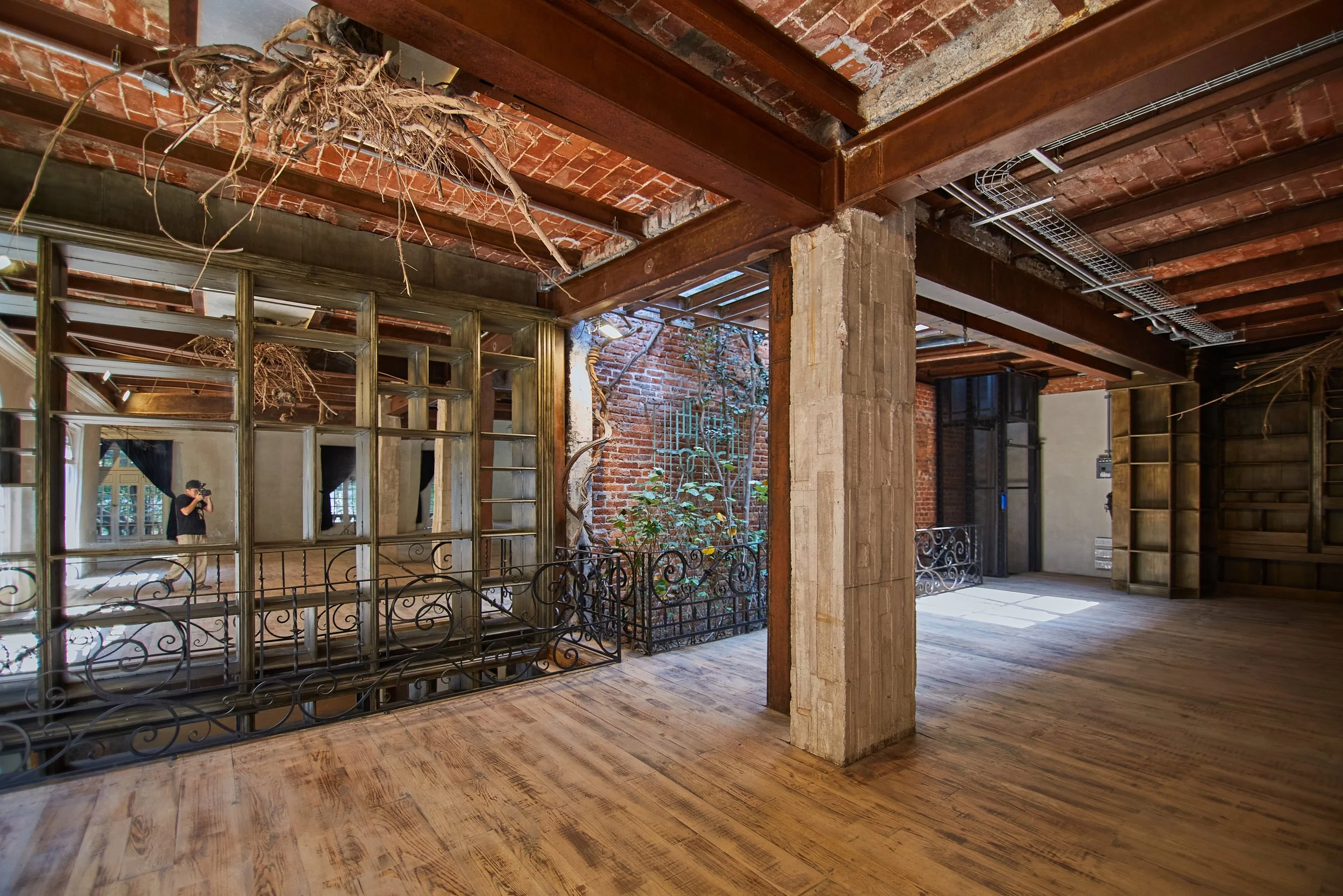 Interior of a renovated industrial-style loft with exposed brick walls, wooden floors, metal beams, and large windows. Some plants and a person taking a photo are visible.