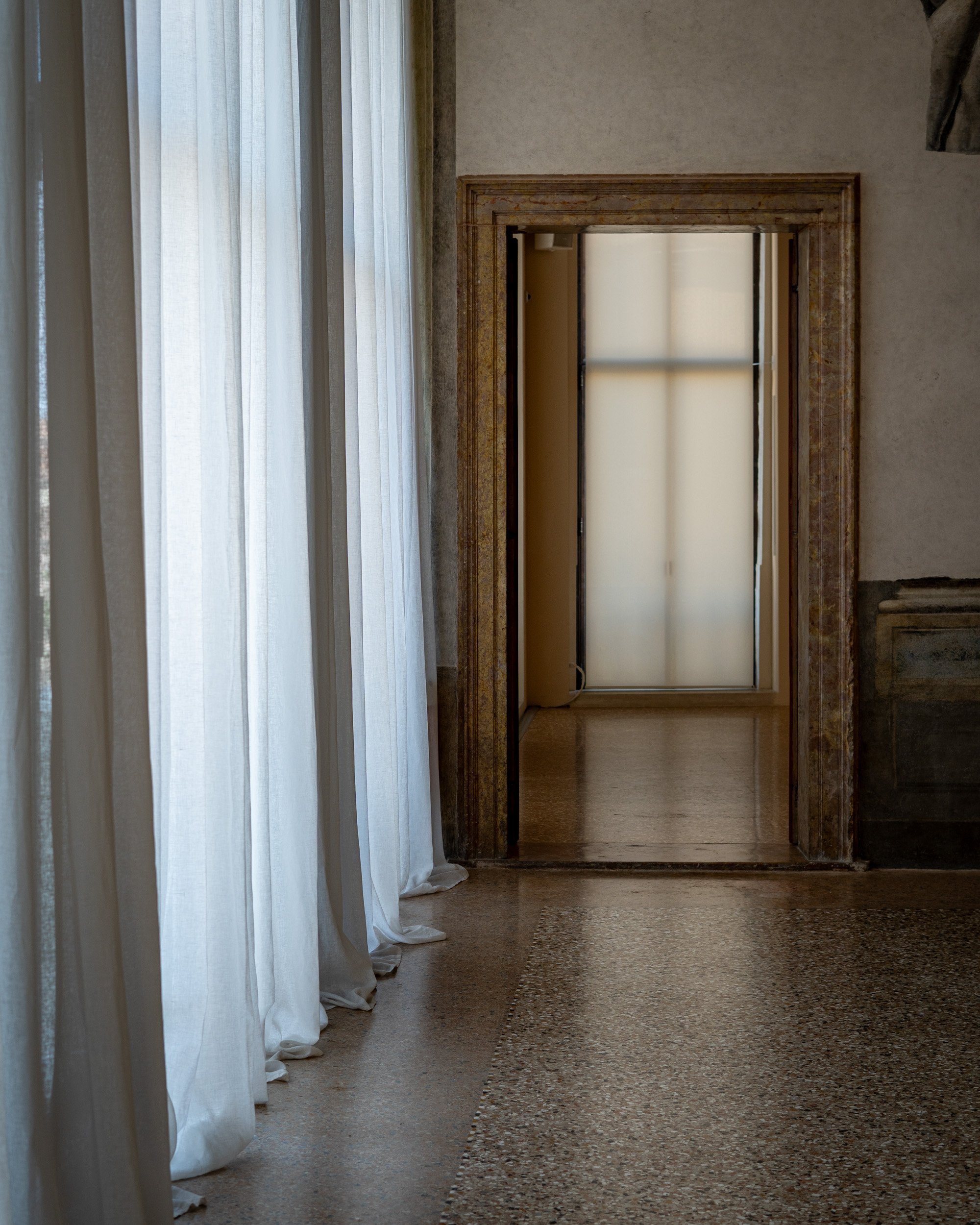 A hallway seen through a doorway with wooden trim. To the left, there are white, sheer curtains hanging from a window. The floor appears to be polished stone or tile with a decorative border.