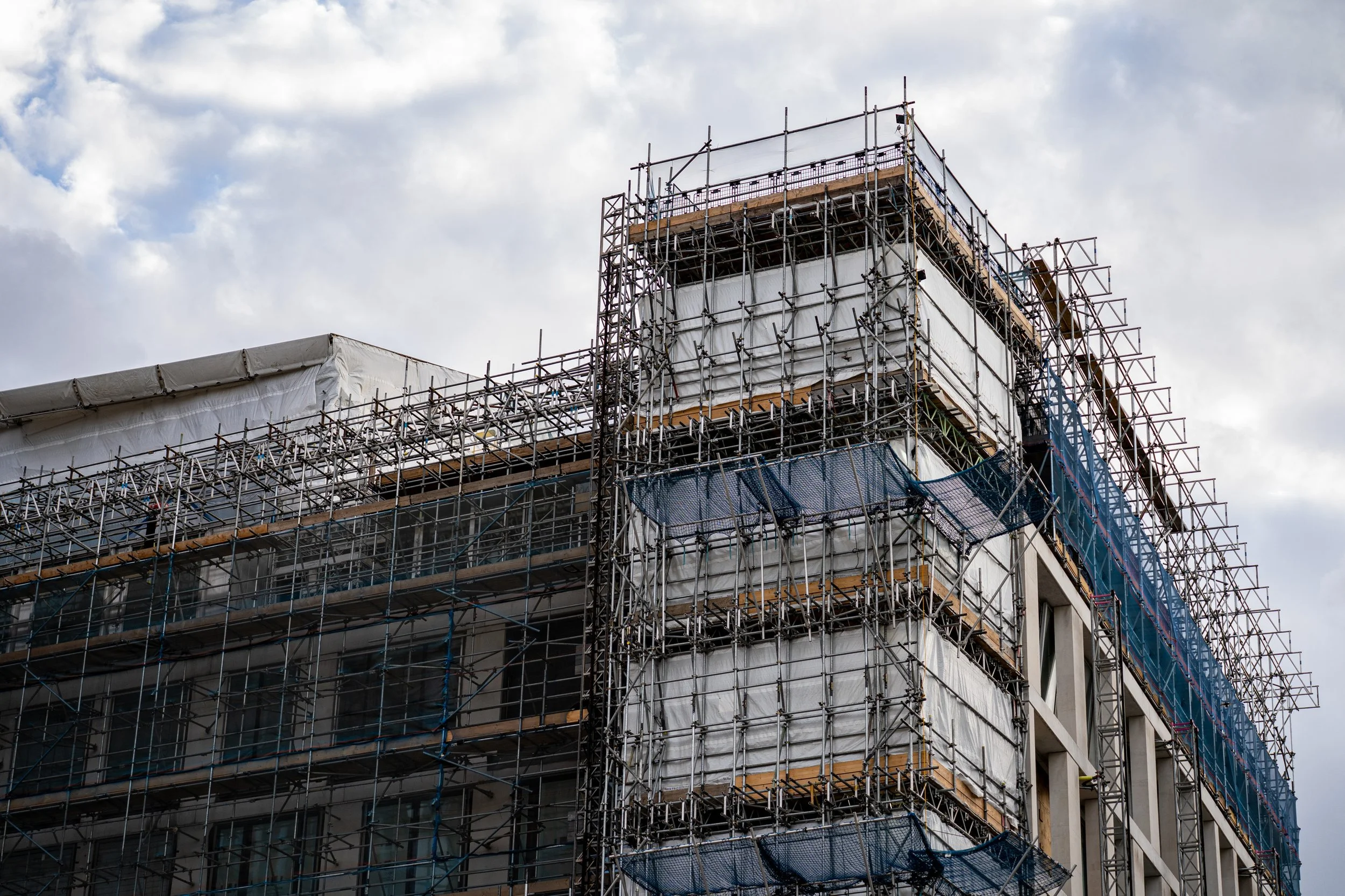 Building under construction with scaffolding and safety netting against a cloudy sky.