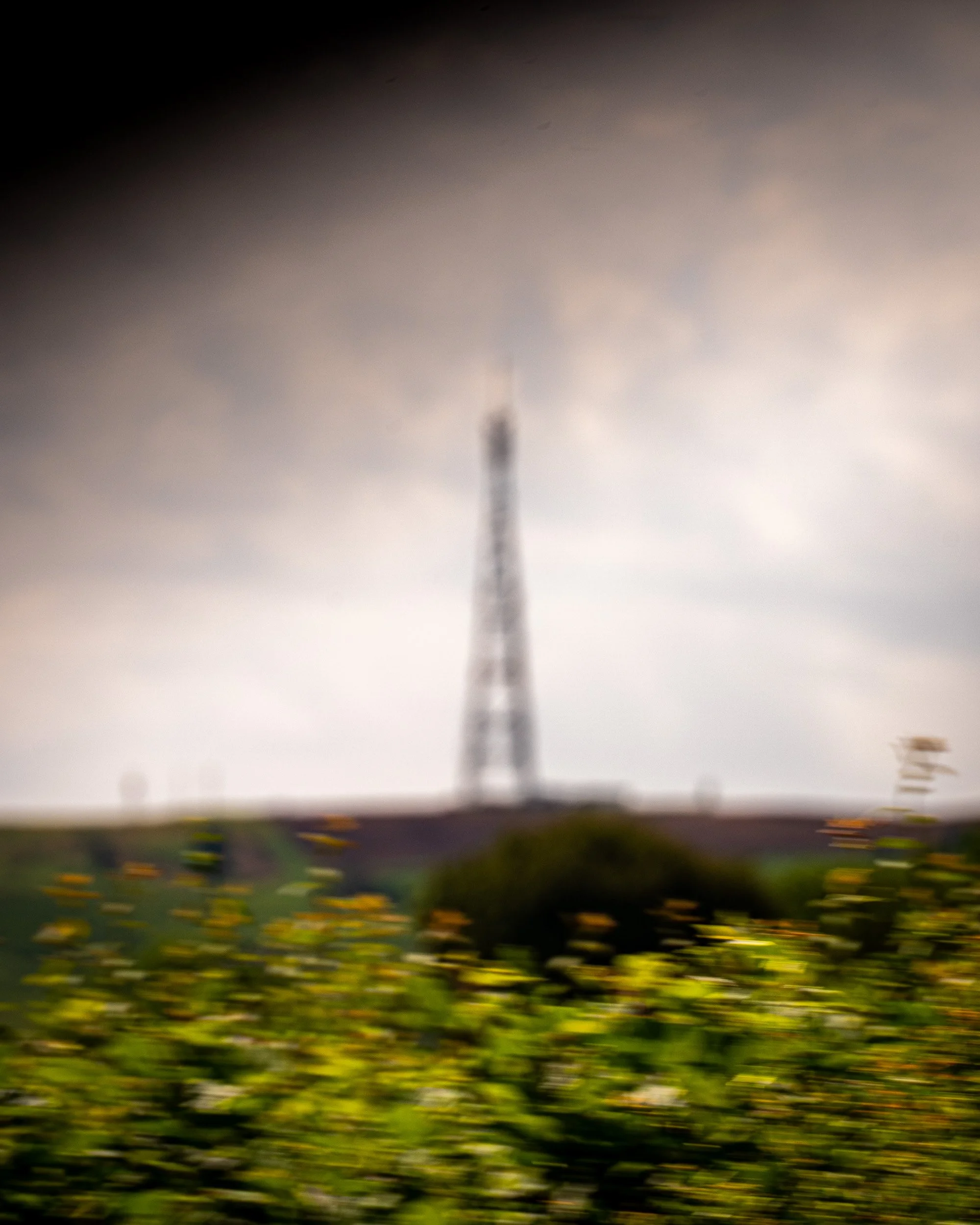 A blurred photograph of a tall tower with a wide base, narrowing towards the top, visible through a dark shadowed frame. The foreground features greenery and yellow flowers.