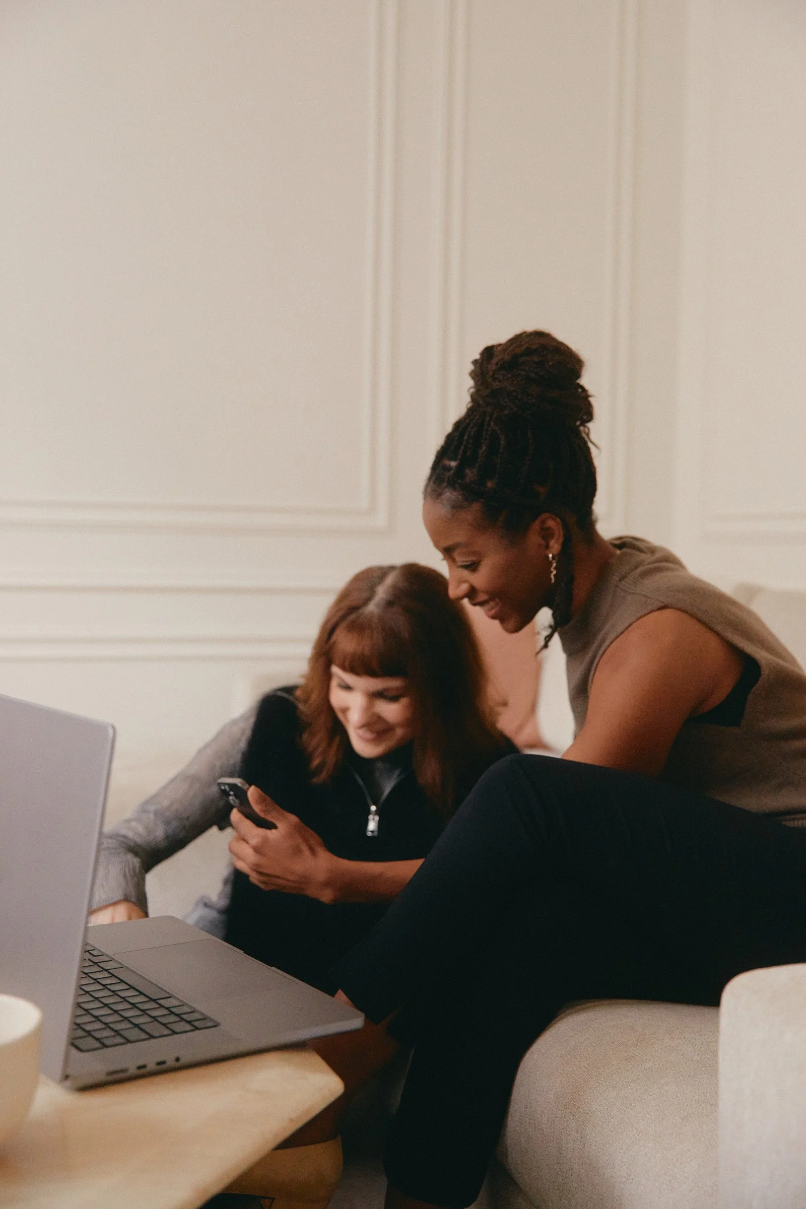 Two women are sitting at a table with a laptop and a smartphone, engaged in a conversation or sharing something on the phone.