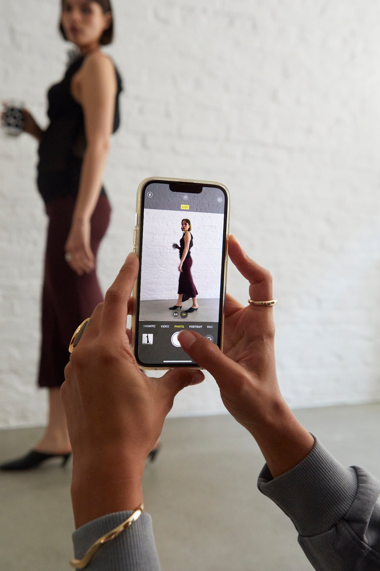 A person holding a smartphone taking a photo of a woman standing in front of a white wall. The woman is wearing a black top and a maroon skirt, holding a cup, and is slightly blurred. The phone screen displays a clear, focused image of the woman in the foreground.
