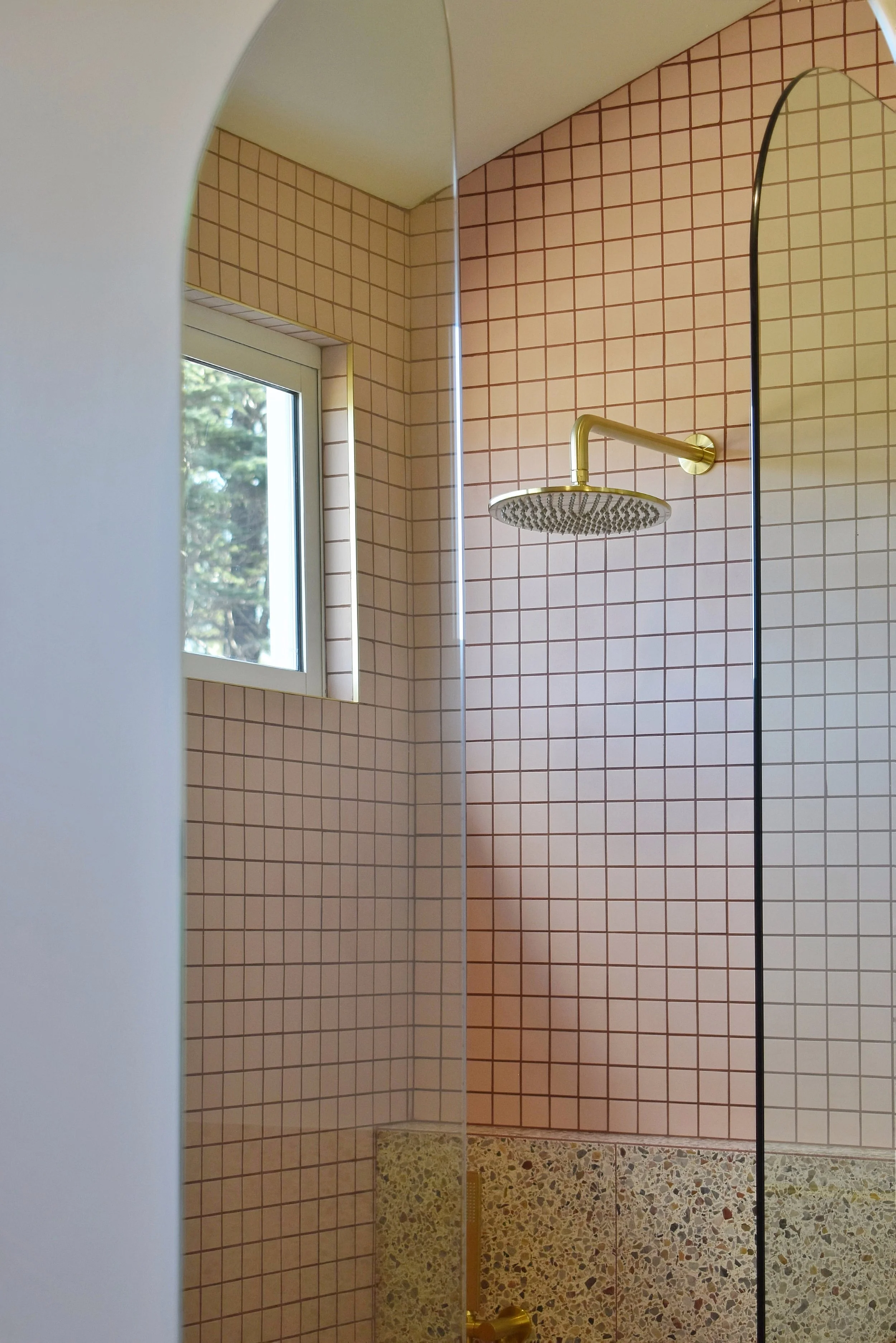 A shower with pink-colored square tiles on the walls, a window showing trees outside, and a brass showerhead mounted on the wall.