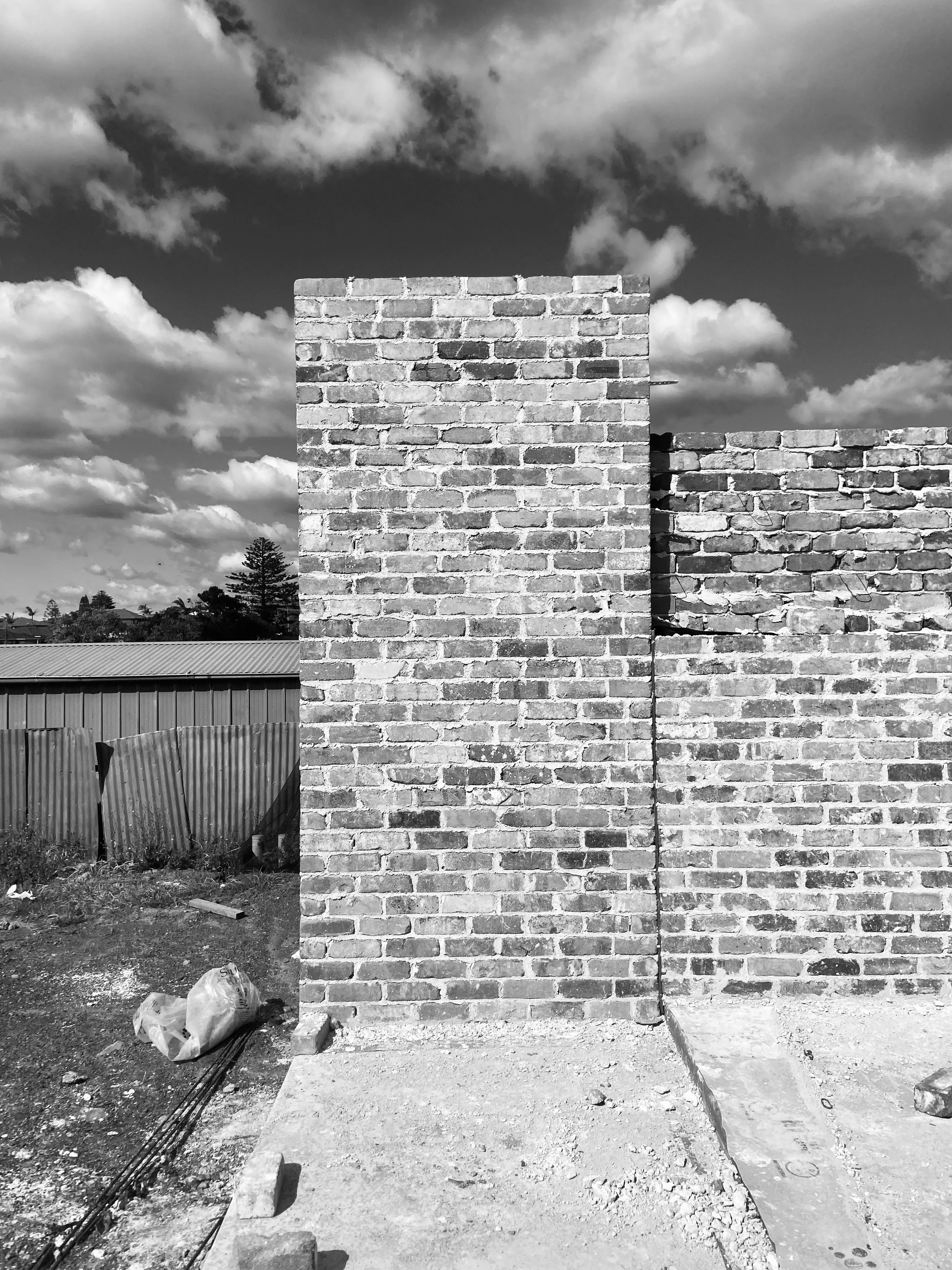 Black and white photo of a construction site with brick party wall and exposed concrete slab on the ground. The background features scenery of the neighbouring coastal properties.