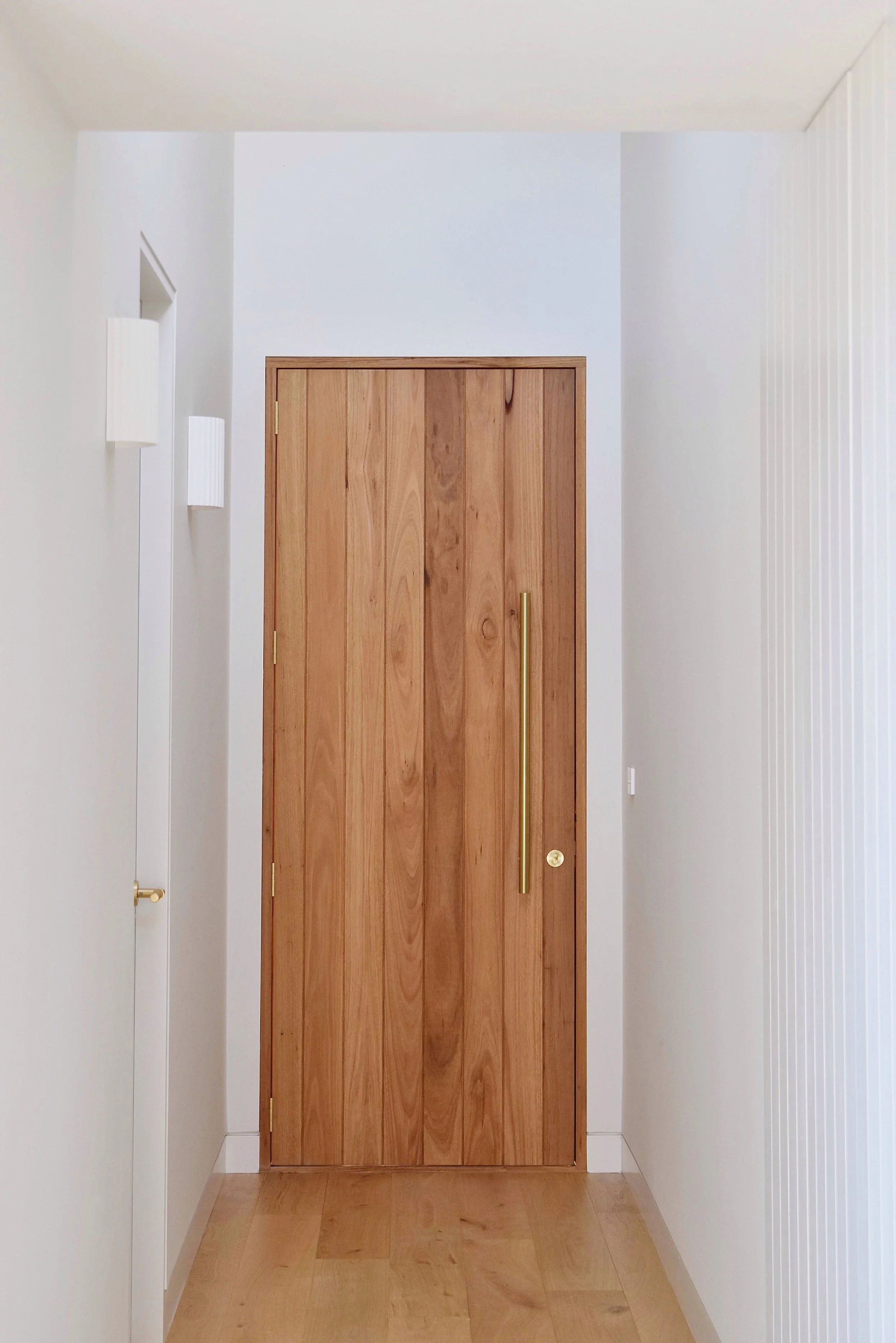 A hallway with white walls, custom timber entry door with brass pull. The floor is light-colored timber, and there is a small brass doorstop on the left wall near the floor. 
