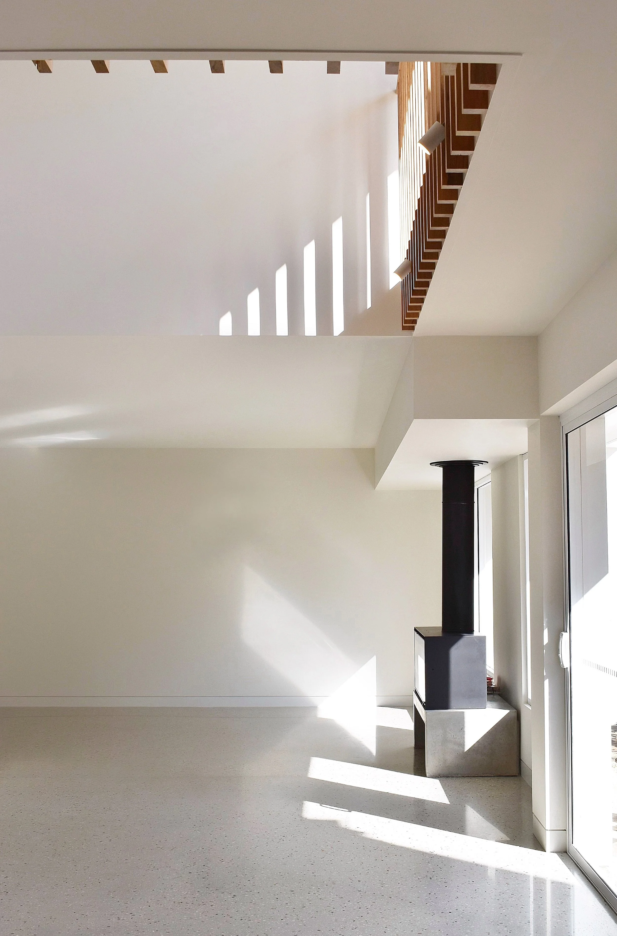 Modern interior space with white walls, wood fire place, large windows, and natural sunlight casting shadows on the floor and walls.