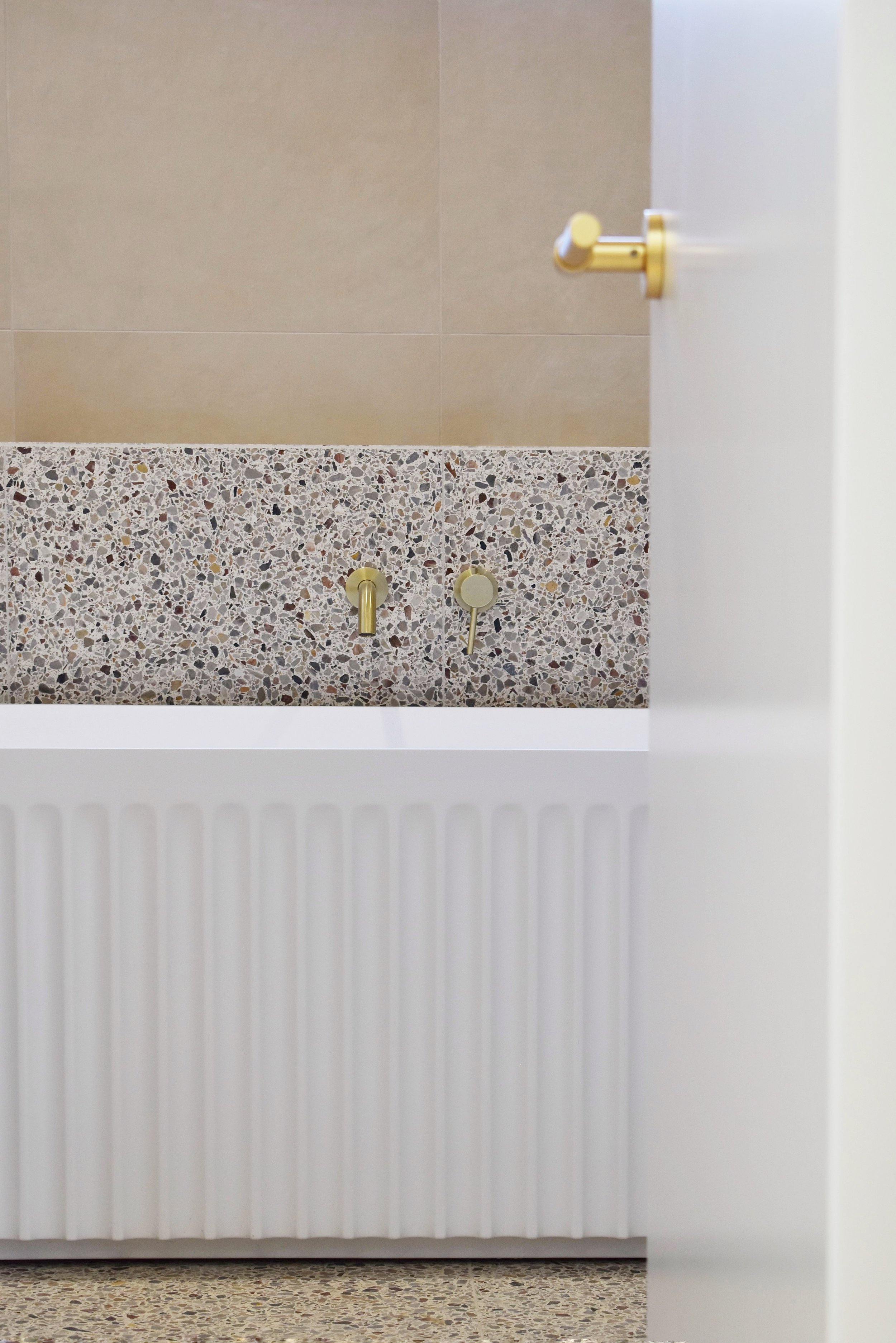 Partially open white door with brass hardware, revealing a beige tiled wall and a speckled terrazzo floor behind it. The bottom part of a white radiator is visible below the door.