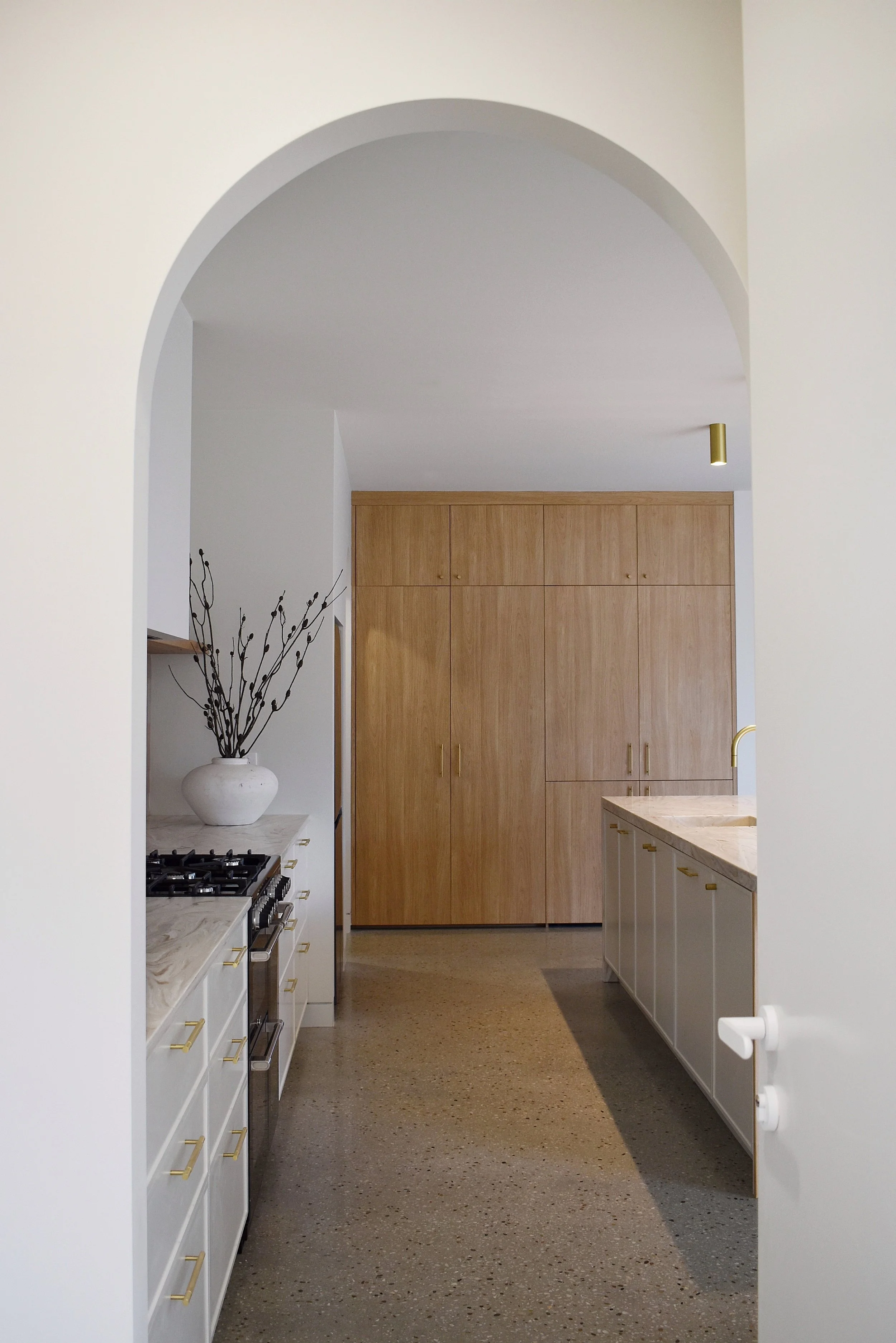 A modern kitchen with white cabinets, gold handles, a marble countertop, and a large wooden pantry. There is a white vase with decorative branches on the counter and a half-height wall with a curved archway.