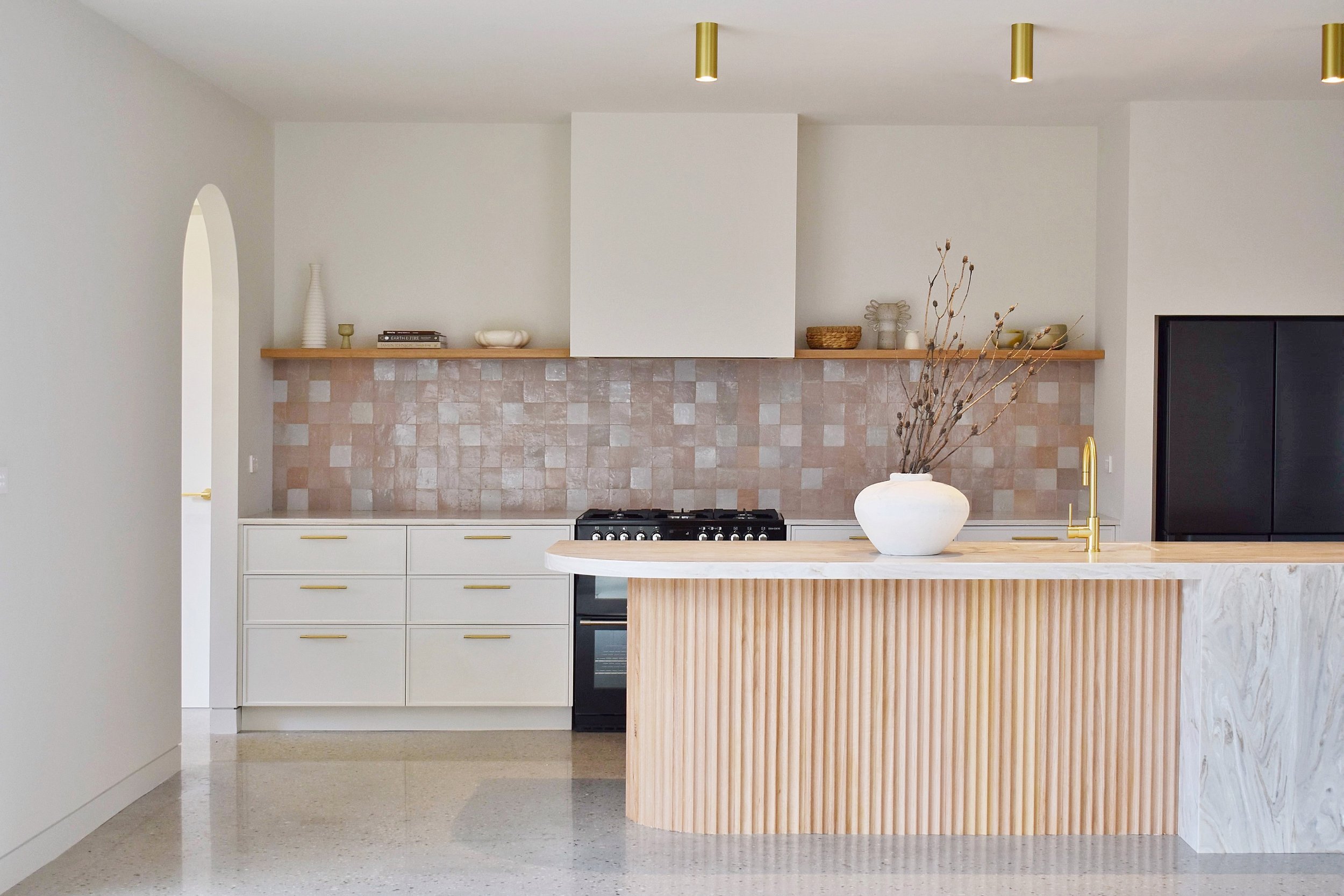 Modern kitchen with white cabinets, a breakfast bar with a wooden ribbed front, a stovetop, and decorative items on a shelf, including vases and bowls, with gold-accented lighting.