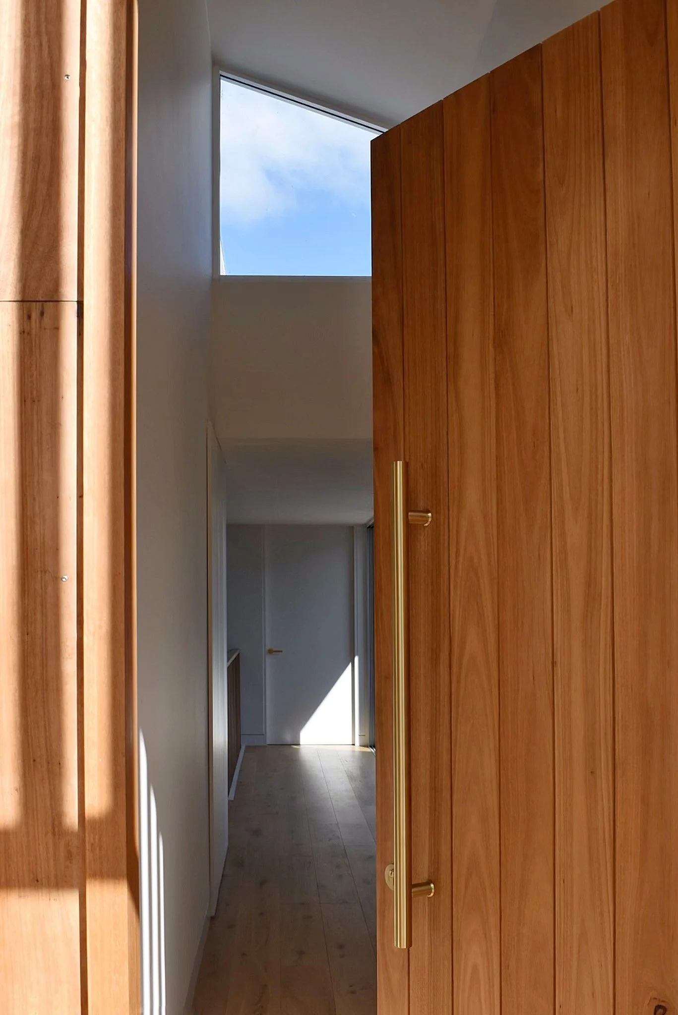 Partial view of a custom timber door with a brass handle, open to reveal a hallway with light-colored walls and a clerestory window showing a blue sky with some clouds.