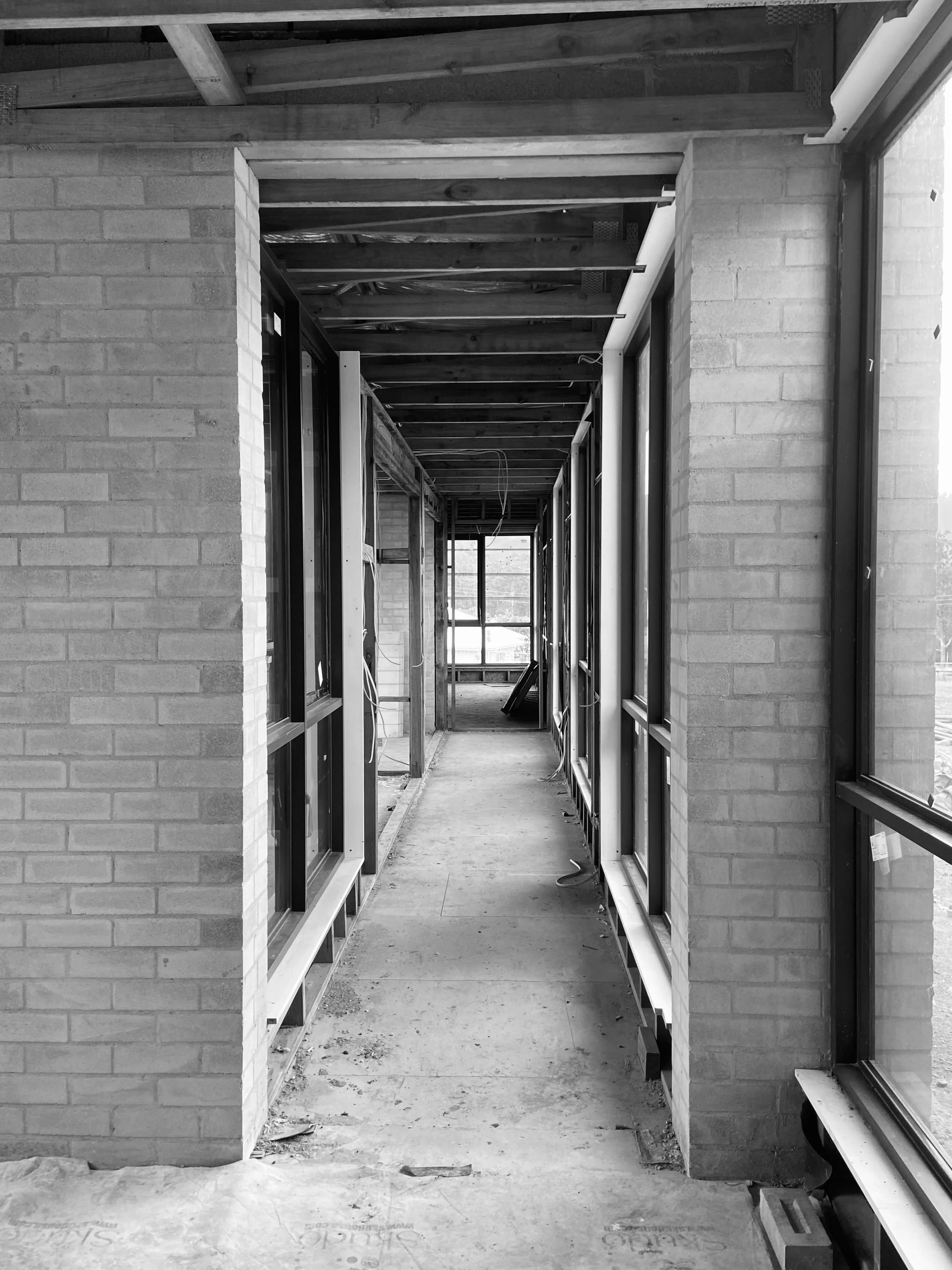 View of a construction site hallway with exposed ceiling beams, window frames, and interior brick walls, showing ongoing building work.