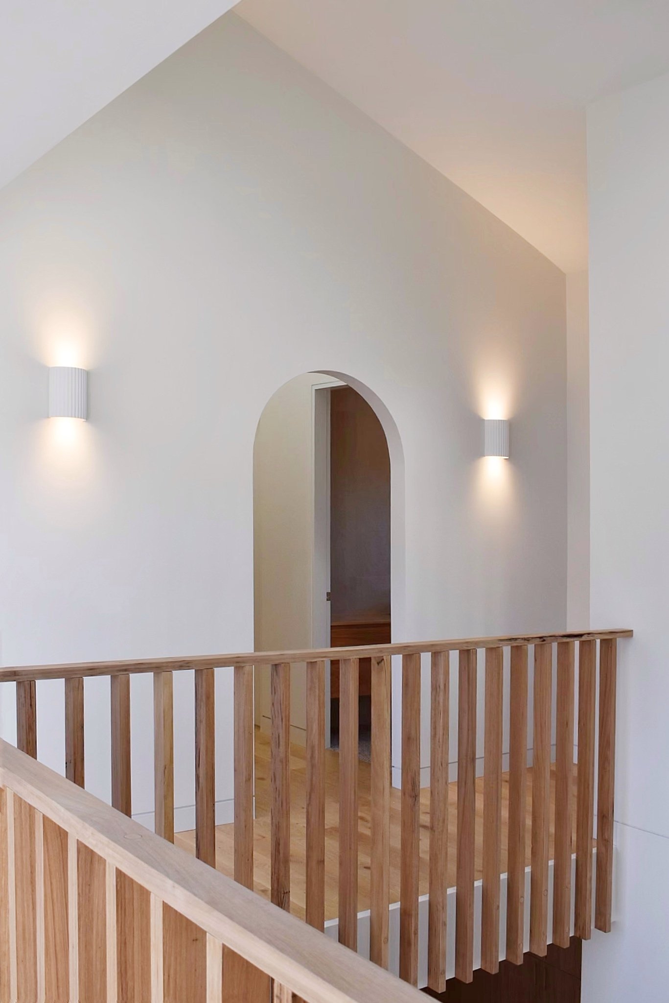 A photo of an interior space with white walls, an arched doorway, two wall-mounted lights, and a timber balustrade.