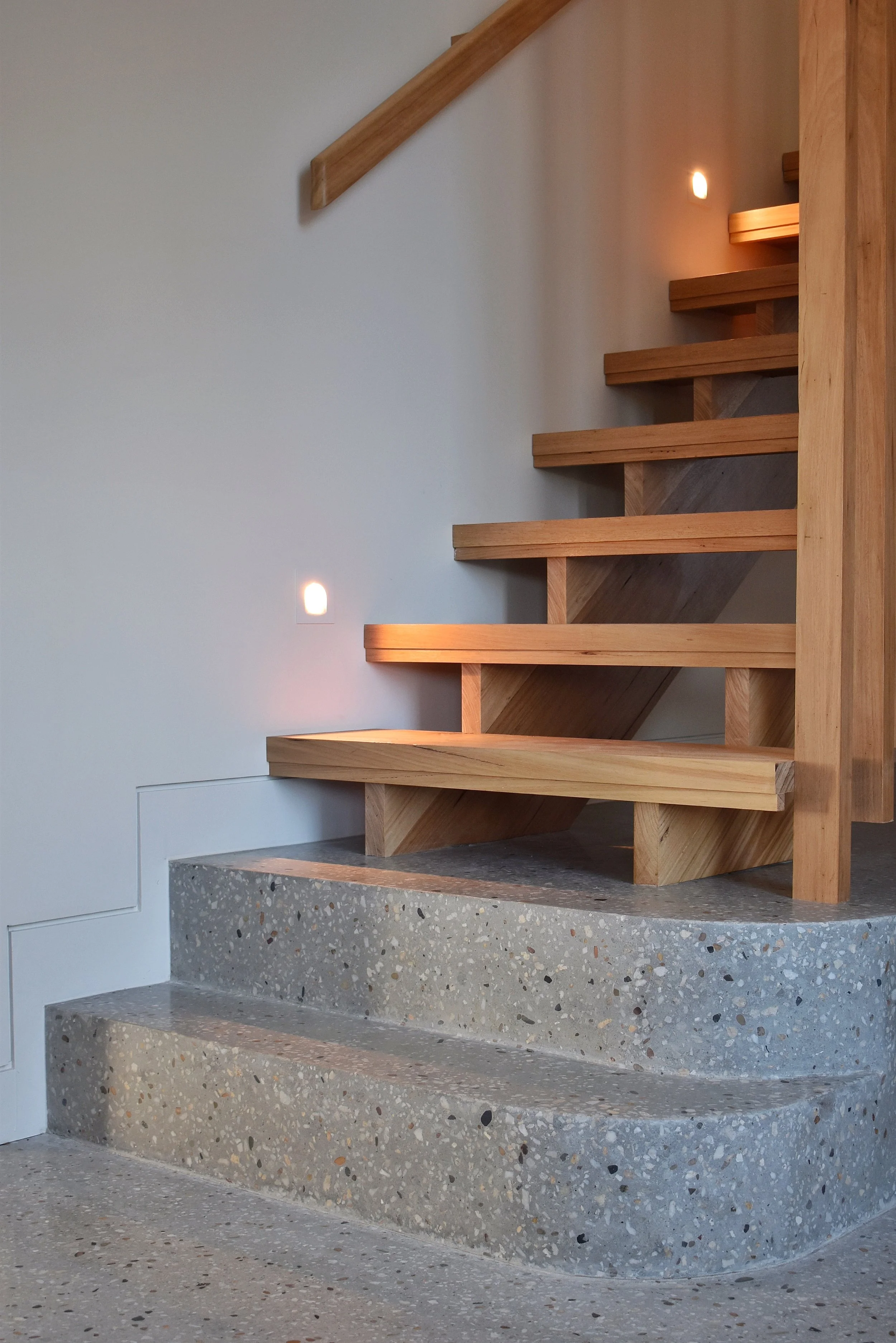 A timber staircase with custom polished concrete curved steps and natural timber handrails and risers illuminated by soft wall lighting.