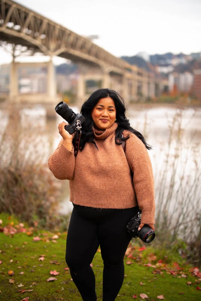 Bethany Villero holds two canon cameras with Portland scenery behind her.