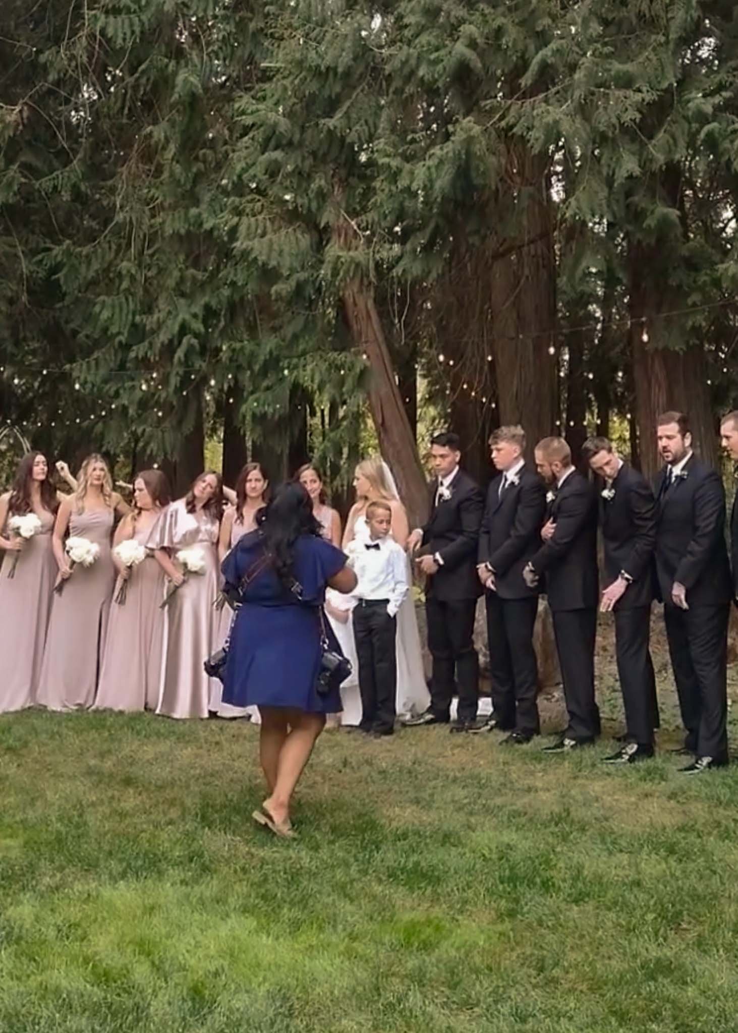A wedding ceremony outdoors with bridesmaids and groomsmen standing in front of large trees, with photographer Bethany Villero dressed in blue.