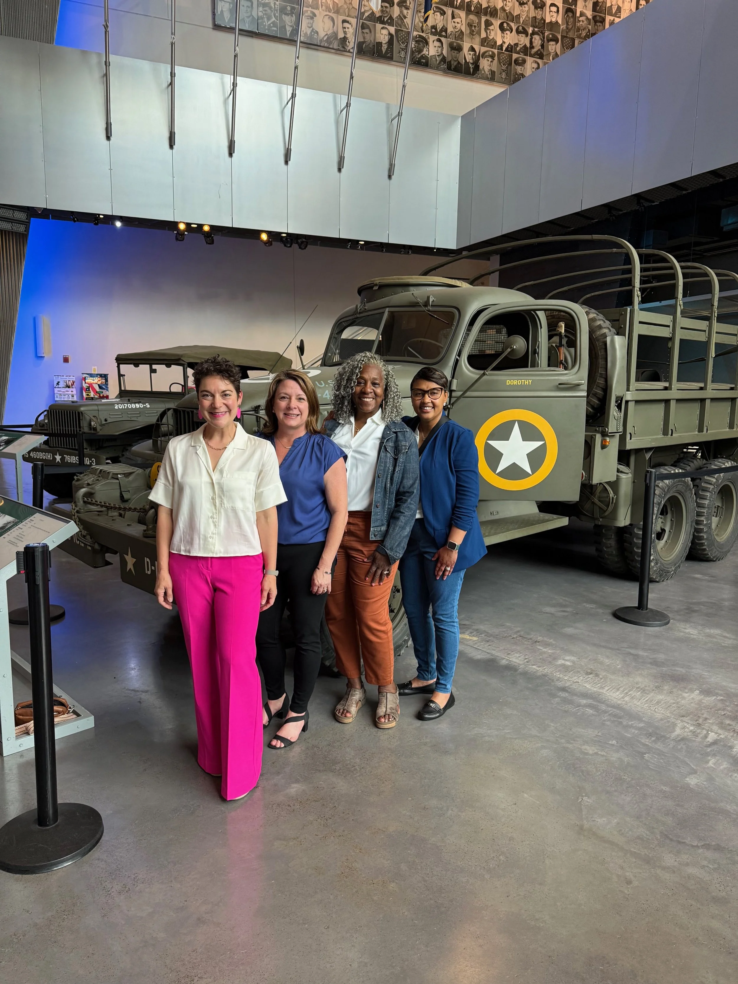 Four women standing together in front of a vintage military truck in a museum exhibit area.