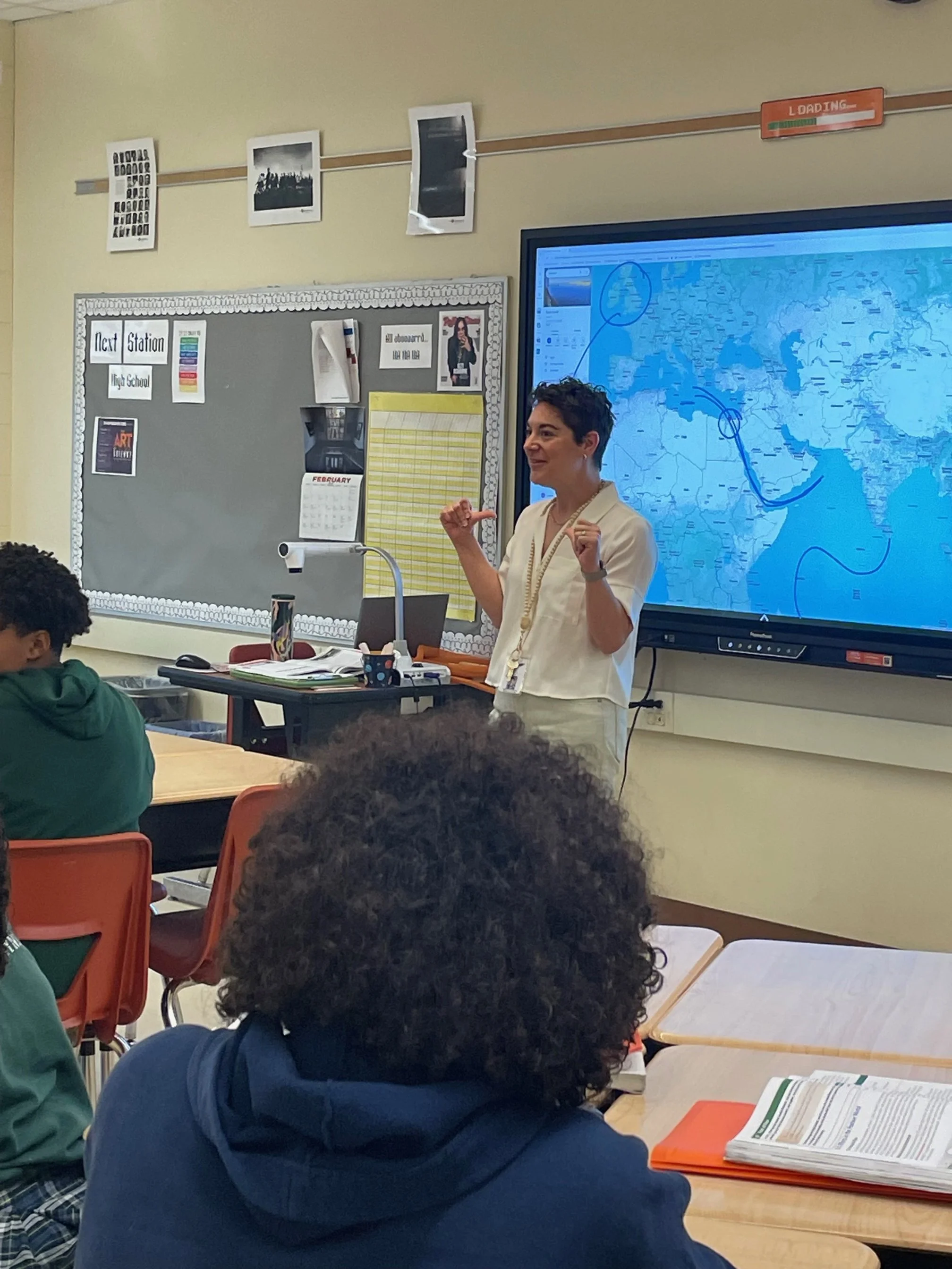 A teacher standing in front of a classroom with students, pointing towards a world map on a large digital screen. The classroom has a bulletin board, photographs, and other educational materials.