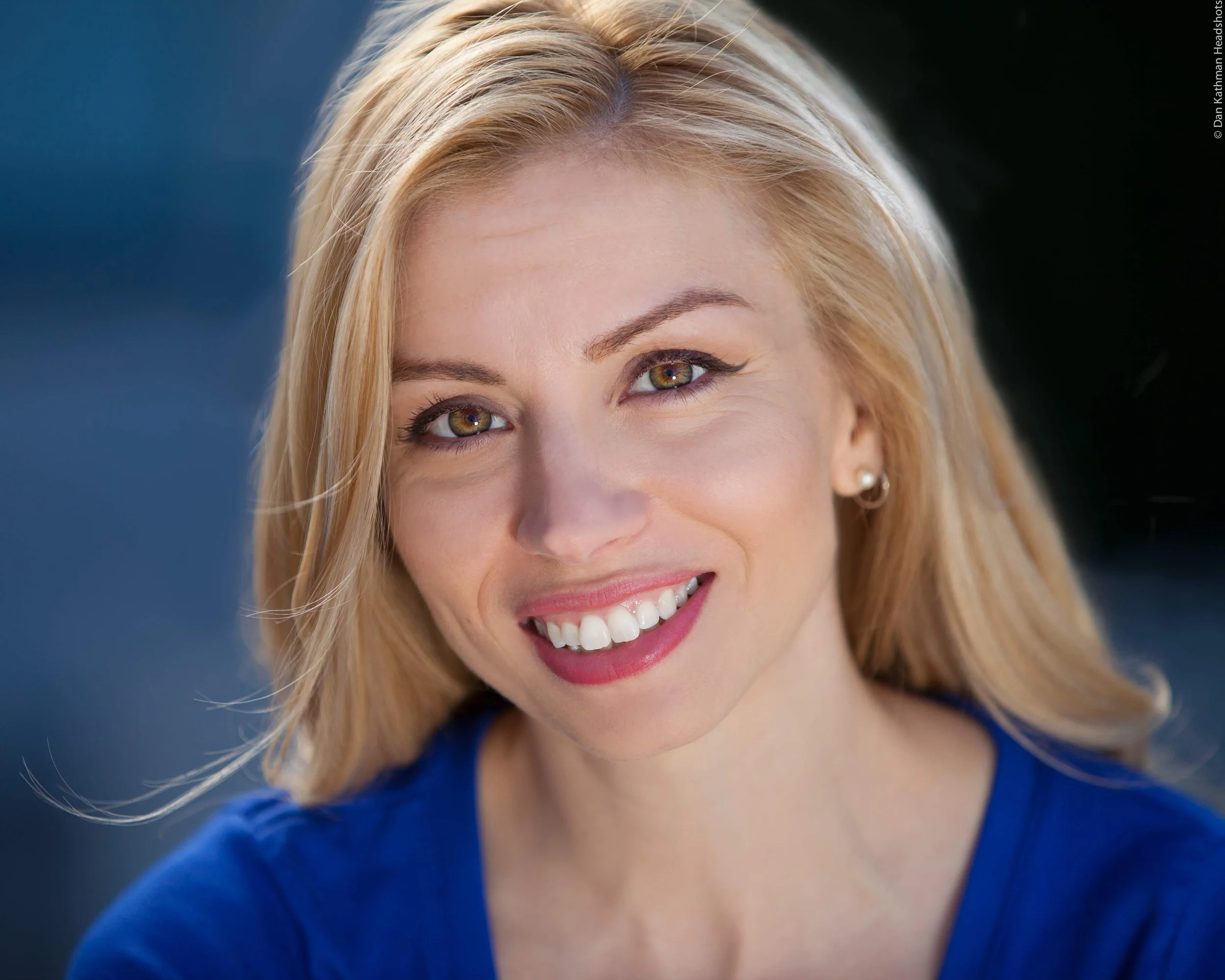 Close-up of a smiling woman with blonde hair, bright eyes, and a blue top, showing her white teeth.