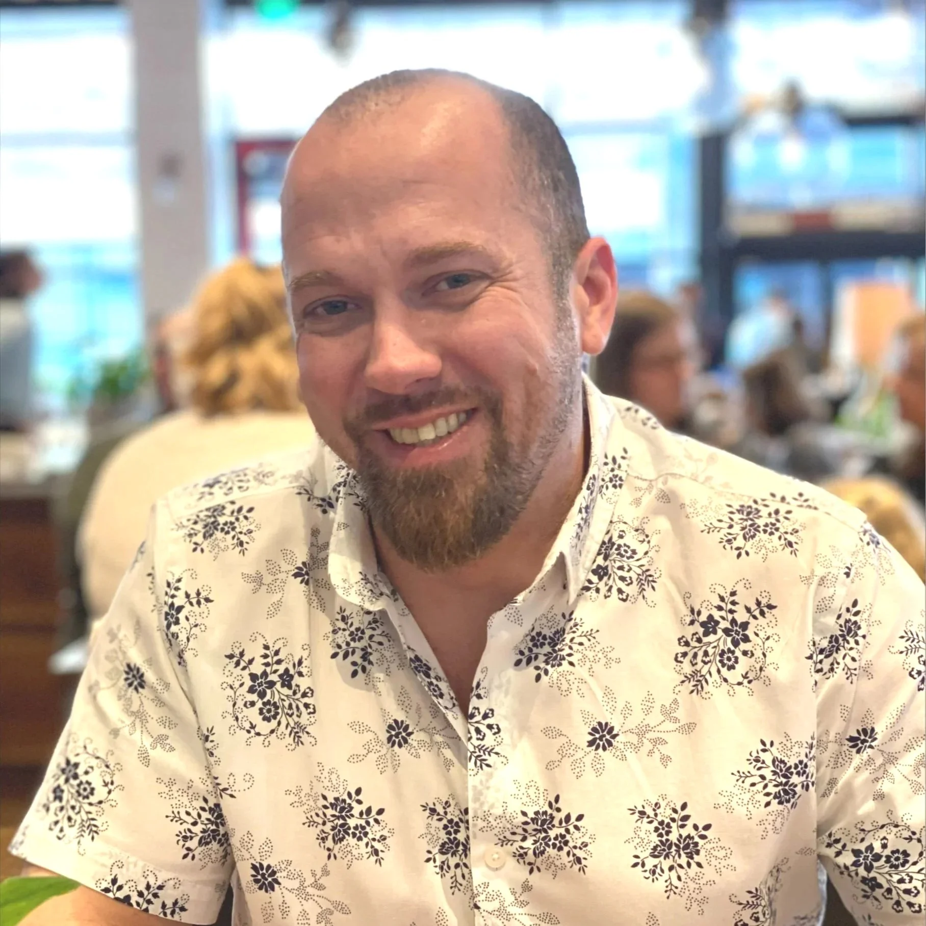 Close-up of a smiling man with a shaved head and beard, wearing a white shirt with black floral patterns, in a busy indoor setting with blurred background of people and windows.