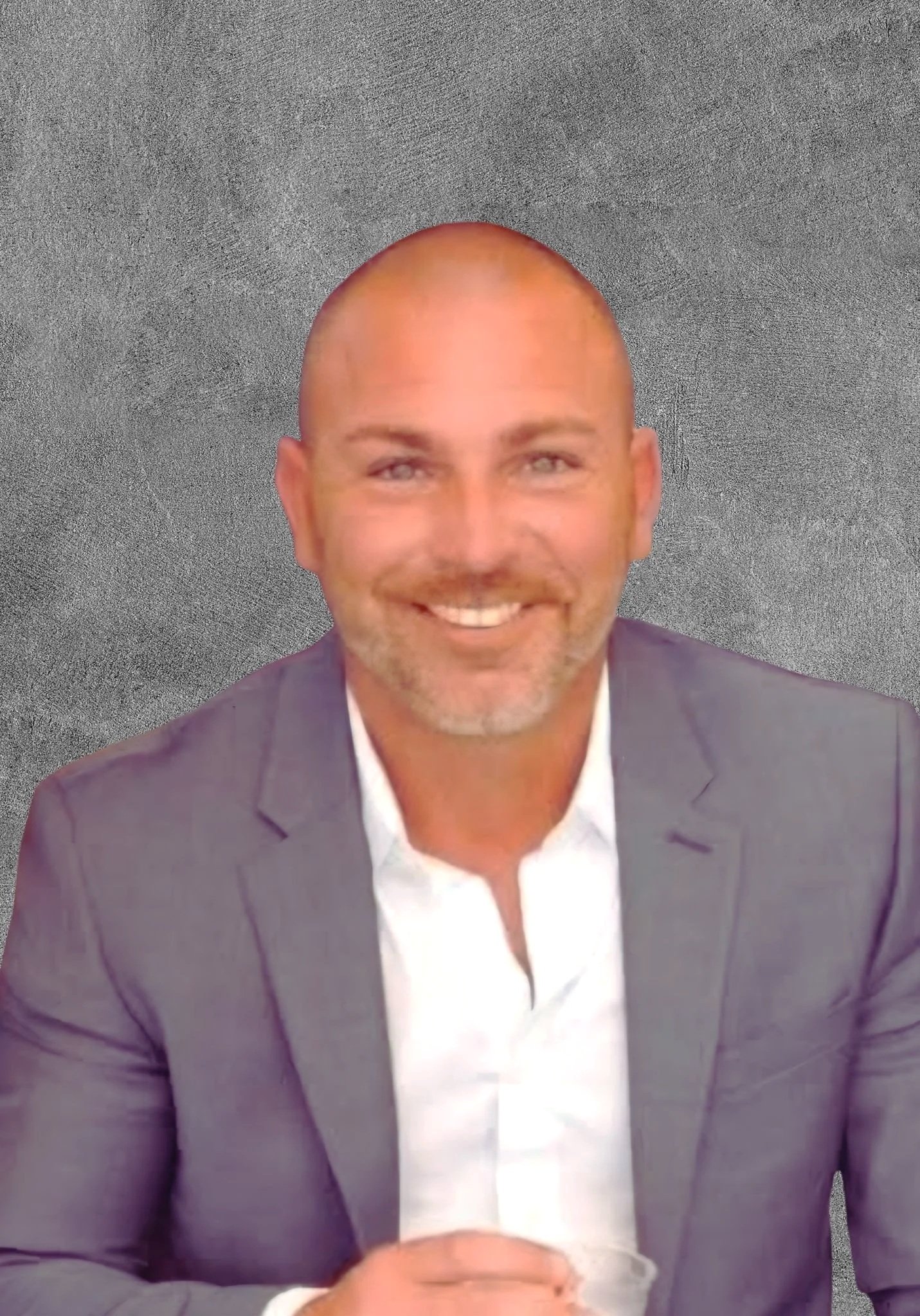 A smiling man with a bald head, dressed in a suit, sitting at a table with a gray textured background.