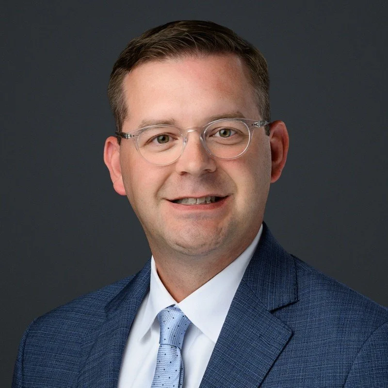 A professional headshot of a man wearing glasses, a dark blue suit, white shirt, and light blue tie, smiling against a dark gray background.